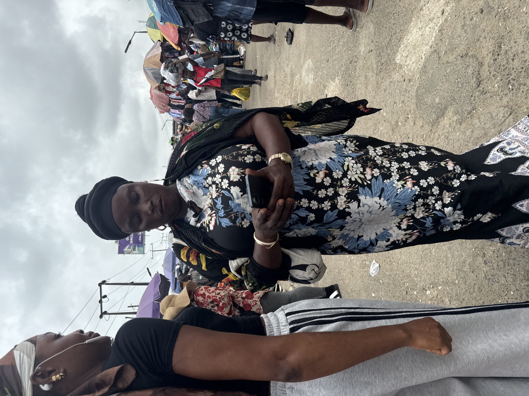 The image captures a bustling outdoor market scene in Lekki, Nigeria, under an overcast sky. In the foreground, a woman wearing a black headscarf and a dark floral-patterned dress is looking down at her smartphone. She is holding the phone in her right hand and has a gold wristwatch on her left wrist. A light-colored handbag with a cartoon character print is slung over her left shoulder. To her left, another woman is partially visible, wearing a black t-shirt and grey sweatpants with black stripes. Her head is wrapped in a brown and white scarf, and she has earbuds in her ears.
The background is filled with various market stalls covered by colorful umbrellas, displaying clothing and other goods. Several other people are visible, some walking and others browsing the merchandise. The ground appears to be a dusty, uneven surface. Electrical poles and wires are visible in the distance, along with streetlights and signage, indicating an urban environment within the city. The overall impression is one of a busy, everyday market day in Lekki.