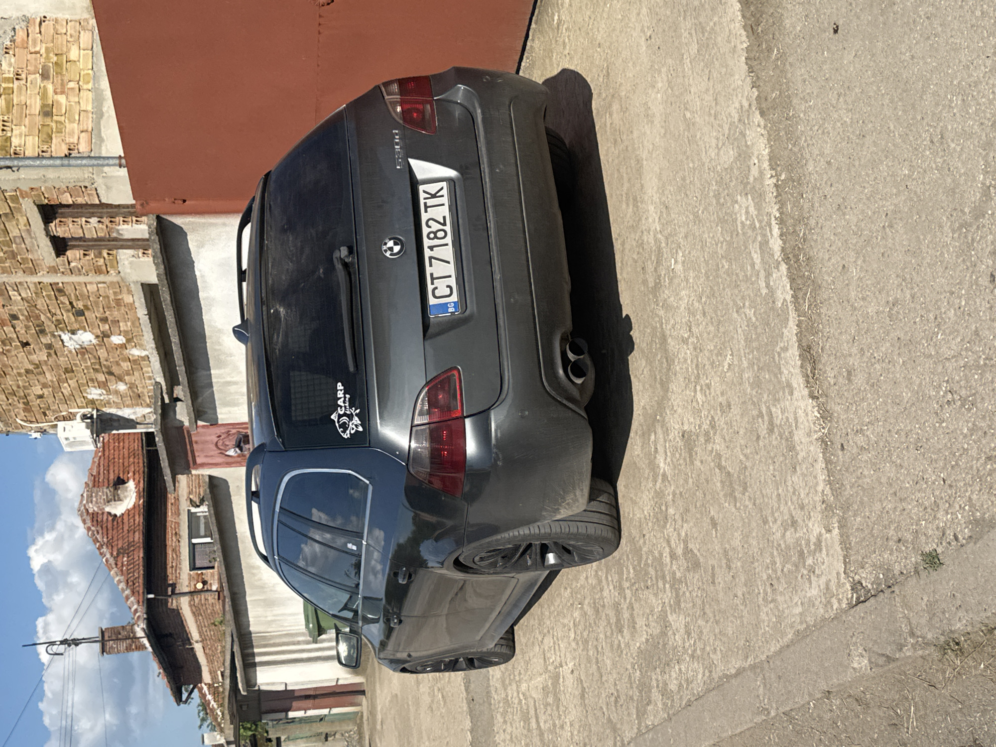 The image depicts a gray BMW 530d parked on a concrete surface. The car has a Romanian license plate number CT 7182 TK. A "Carp Fishing" sticker is visible on the rear window. The background includes a mix of buildings, some brick and some with red roofs, characteristic of an urban or residential setting. Based on the location provided, the image is likely taken in or near Sarnevo, Bulgaria. There are no people visible in the image. The car appears to be stationary. The environment appears to be outdoors on a sunny day.