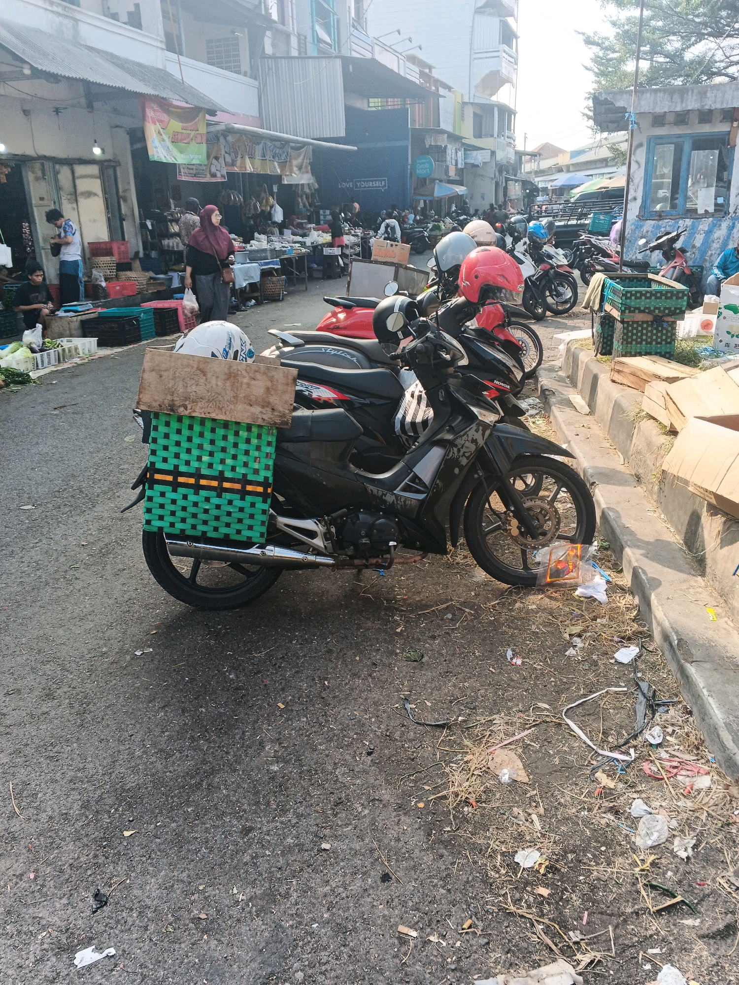 The image depicts a street scene in Ungaran, Indonesia, likely a market area. The foreground is dominated by several parked motorcycles. The most prominent motorcycle has a green woven basket with orange accents attached to the rear, holding a wooden plank and a white helmet. Other motorcycles are visible, some with red and black accents, and one with a red helmet. In the background, a row of market stalls lines the street, with awnings overhead. People are seen browsing or working at the stalls, with one woman in a red hijab walking with a plastic bag. The ground is asphalt, with scattered debris including plastic wrappers, straw, and other litter. The environment suggests an active marketplace with a mix of commerce and transportation.