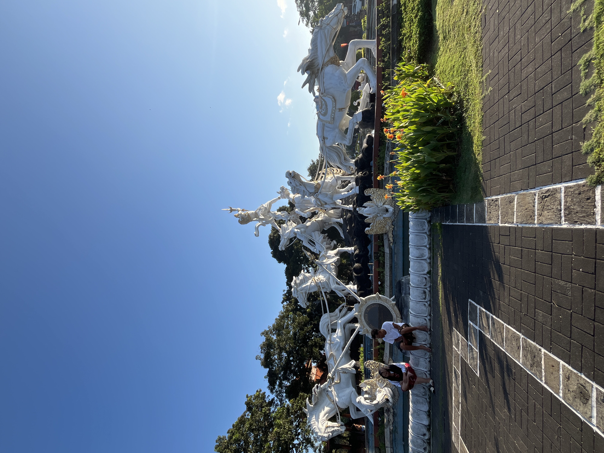 This image captures a wide shot of the Grand Dewa Ruci statue in Kuta, Bali, Indonesia, a prominent landmark depicting a mythological warrior. The statue, predominantly white, features a chariot pulled by multiple horses, with a warrior figure at the helm. In the foreground, two individuals are seated on a stone ledge. One is a female wearing a headscarf and a red and white top with a red skirt, and the other is a male in a white t-shirt and dark shorts. They are positioned to the left of the statue. The statue complex is surrounded by lush greenery and decorative plants with orange flowers. The ground in the foreground is paved with dark rectangular bricks, with white painted lines forming a hopscotch-like pattern. The sky is a clear, bright blue. The lighting suggests it is either early morning or late afternoon, with the sun casting shadows.