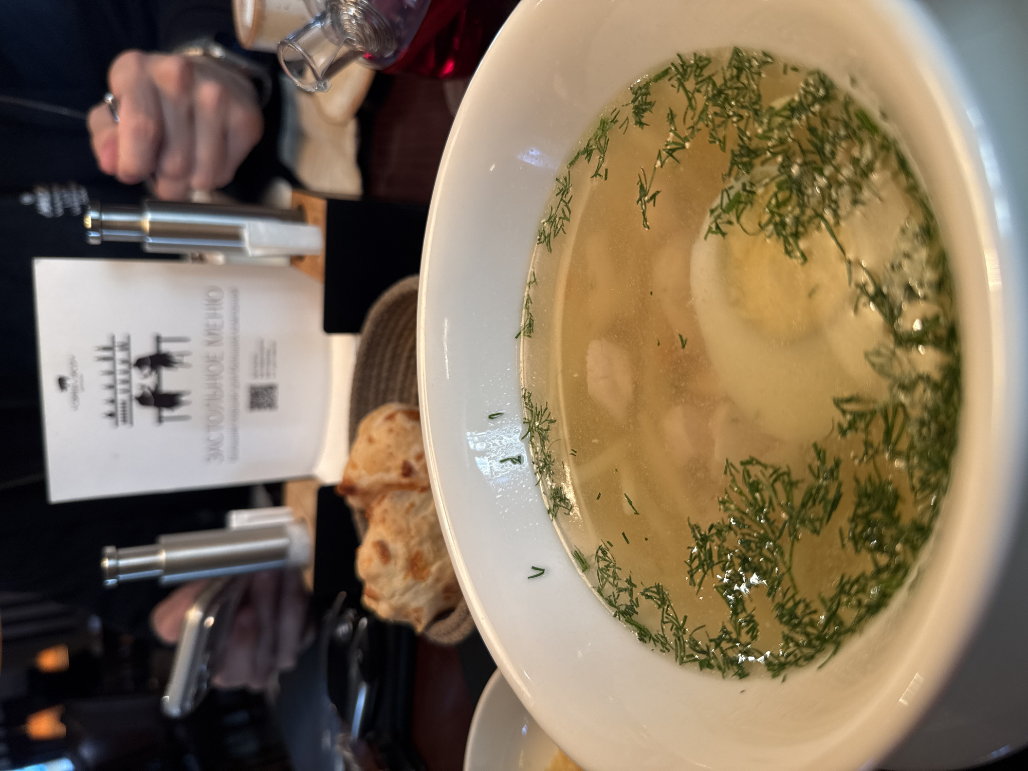 A close-up shows a bowl of chicken soup with dill, an egg, and pieces of chicken, positioned on a table in an indoor setting. Next to the soup is a basket of bread rolls, a menu card with Russian text (“Застольное меню”) and a QR code, and two silver salt and pepper shakers. A person's hand wearing a watch is visible on the left side of the frame. The location is Krasnoyarsk, Russia.