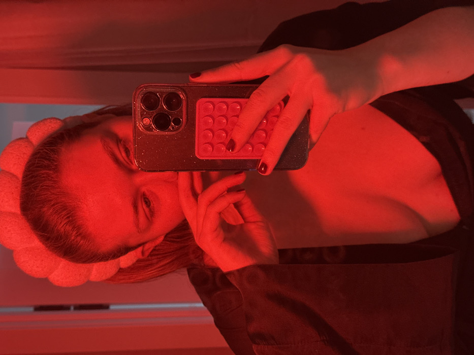 This image is a mirror selfie taken indoors in Nikol'skii, Russia, under strong red lighting. The central subject is an individual with hair pulled back, wearing a light-colored, segmented headband and a dark, open top or robe that reveals a portion of their upper chest. A dark strap with white markings is visible on the left side of the chest area. The subject's right hand holds a dark-colored smartphone with a glittery case and a triple-lens camera module. Attached to the back of the phone is a red, rectangular accessory featuring a grid of circular indentations. The subject's left hand is positioned near their lower face. Fingernails on both visible hands are painted dark. The individual is looking downwards, likely at the phone screen. The background is largely obscured by the red light, showing only faint reflections and a vertical line suggesting an architectural element like a doorframe.
