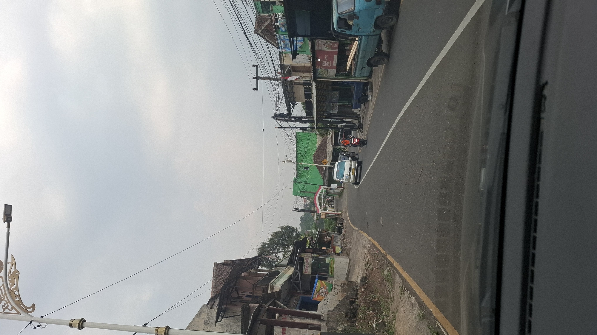 The image depicts a street scene in Cilaku, Indonesia, on an overcast day. A paved road runs through the center of the frame, with buildings lining both sides. On the left, a two-story structure with a weathered facade and a red tile roof is visible, featuring open balconies and shops at ground level. A motorcycle is parked in front of one of these shops. Further down the road, a white van is in motion, and a woman on a motorcycle, wearing an orange hijab, is also visible. Across the street, a vibrant green building stands out, with an archway adorned with the Indonesian flag. To the right of the road, a light blue pickup truck is parked, loaded with lumber. A power line pole with multiple wires stretches across the upper portion of the image, indicating the presence of electrical infrastructure. The overall environment suggests a semi-urban or rural area with a mix of commercial and residential buildings.