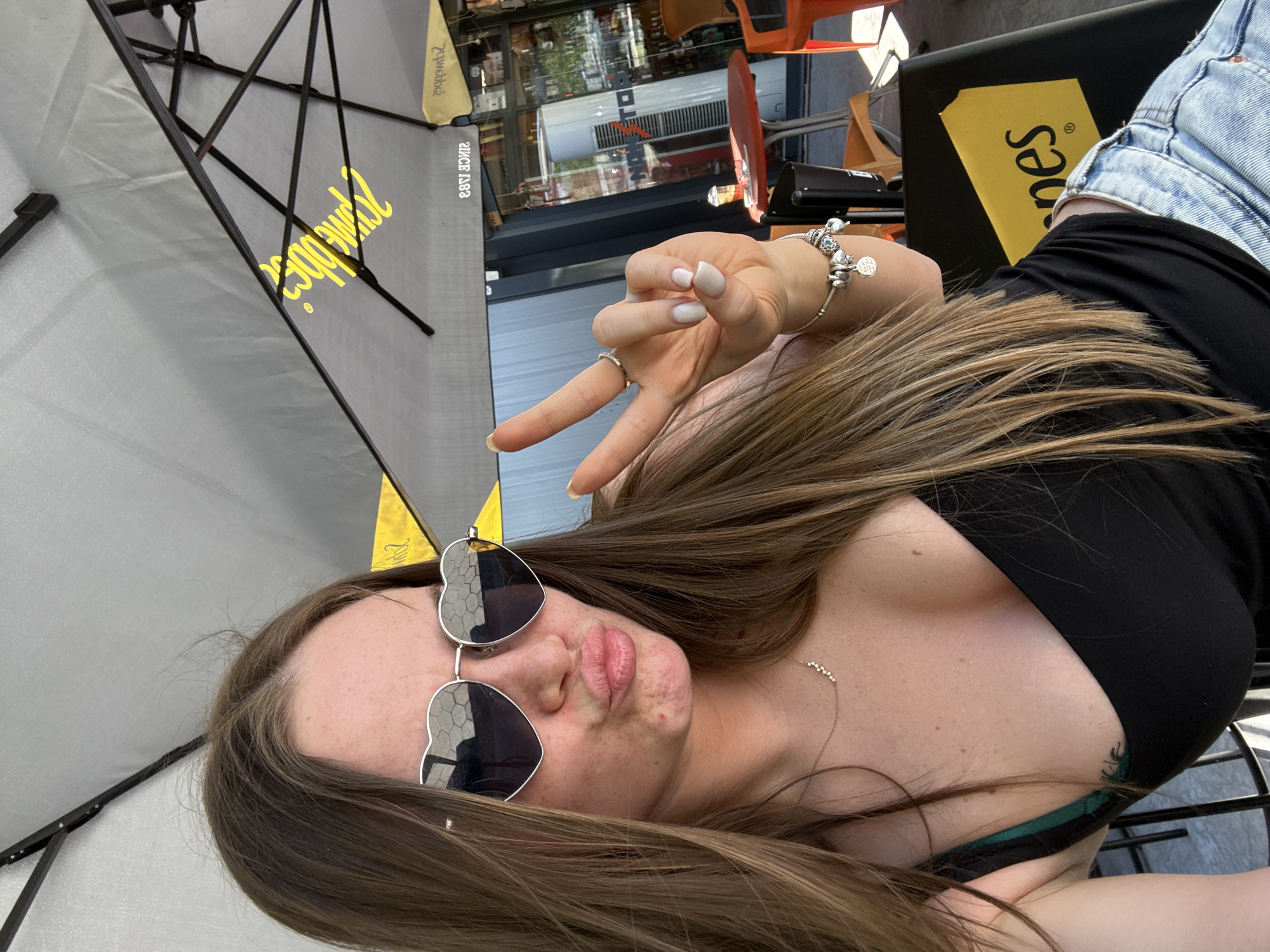 A medium close-up depicts a young woman posing outdoors. She is wearing heart-shaped sunglasses and a black top, showing a hint of a green bra strap. A silver necklace is visible on her chest. She is making a peace sign with her hand, which has a silver ring and a charm bracelet. She is wearing denim shorts. The background shows a partially visible umbrella, a commercial storefront with reflections, and outdoor seating, suggesting a cafe or restaurant. The outdoor seating includes orange chairs and black furniture with a yellow sticker with the word "Jones" written on it. The scene appears to be in the city of Plovdiv, Bulgaria.