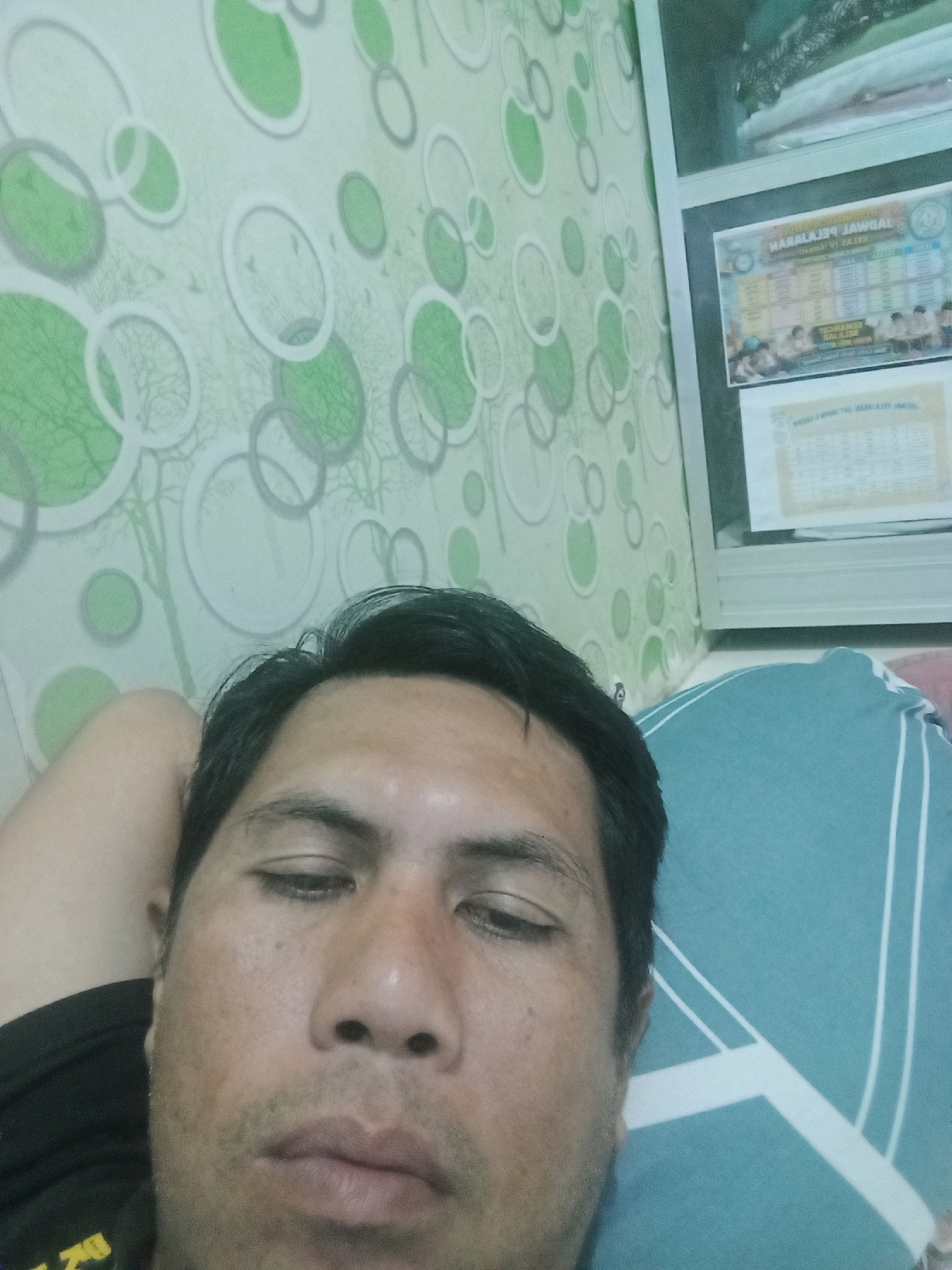 The image is a selfie taken indoors. A man is lying down with his head propped on his arm, looking towards the camera. He has dark hair and is wearing a black t-shirt. The background consists of a wall with a green and white circular pattern wallpaper and a white shelf unit with some papers and what appear to be folded clothes. The scene appears to be a bedroom. The location is stated as Ketapang Lor, Indonesia.
