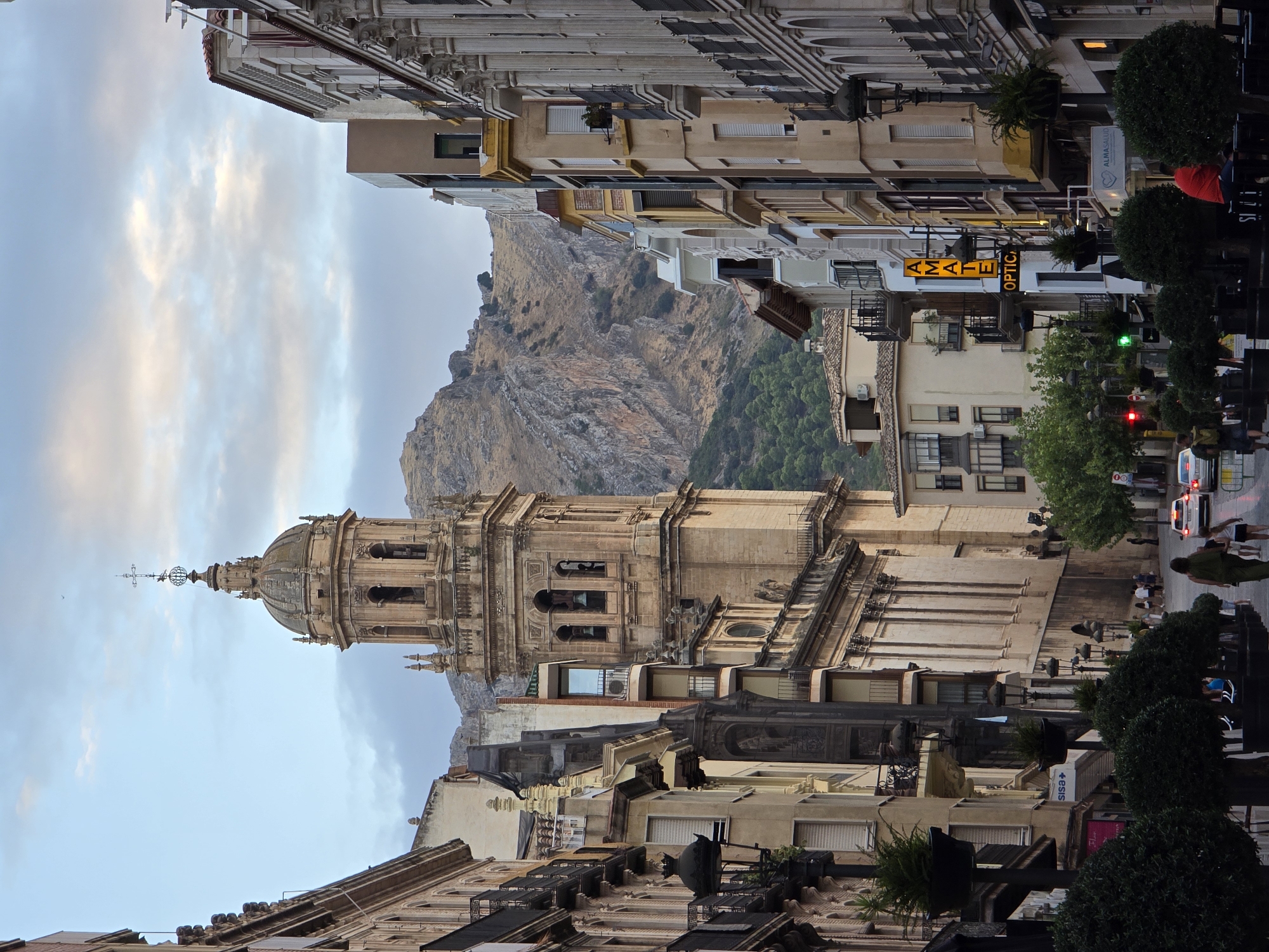 The image captures a view looking up a wide, cobblestone street in Jaén, Spain, towards a prominent bell tower of a cathedral. The architecture is colonial with a prominent church structure on the right and other buildings lining the street on both sides. The buildings have balconies and ornate facades. A mountain range is visible in the background.
There are several people visible on the street. A woman in a green dress and a girl in a white top and dark shorts are walking away from the camera. Further down the street, several people are walking and gathered in small groups. Two white cars are on the street, one of which appears to be a police car due to its flashing blue lights. A man in a red shirt is sitting on a bench.
The street is lined with trees in large planters, and there are several lampposts. Signs for businesses are visible, including "AMATE OPTICA" and "ALMASALUD". The sky is partly cloudy with soft light, suggesting it is either early morning or late afternoon. The overall scene depicts a vibrant urban environment with a blend of historic and modern elements.