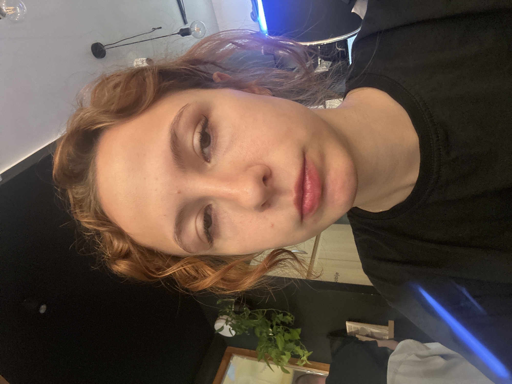A close-up, rotated portrait shows a fair-skinned person with reddish-brown, curly hair, a black shirt, and lightly tinted lips. The background includes a blurred view of a black surface, a plant, and partial reflections in a mirror, likely within an indoor setting. Based on the text "oblije" visible on an object, it's possible that the location is in Novi Sad, Serbia. The environment appears to be lit by a mixture of artificial and natural light.