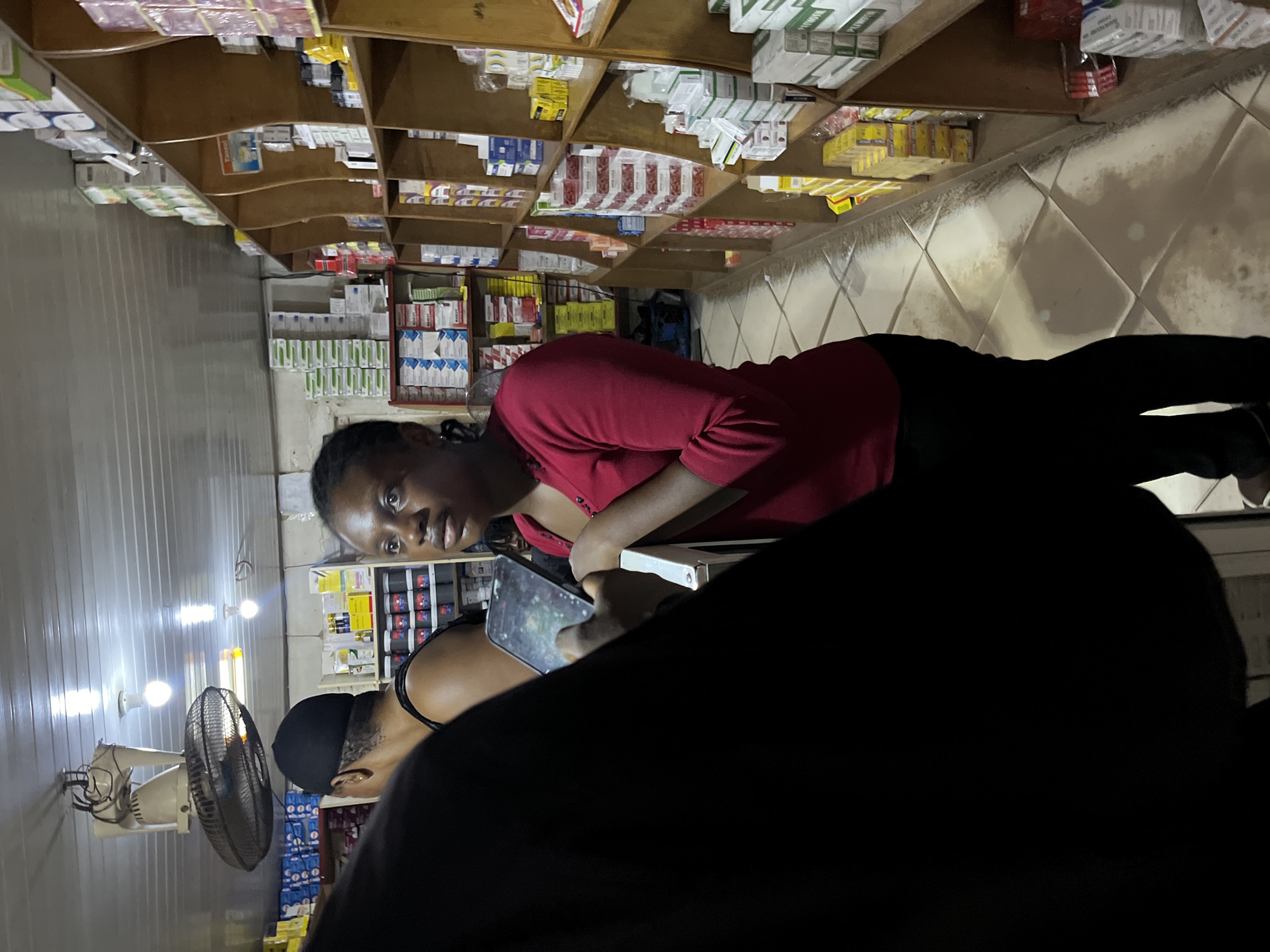 In Bukuru, Nigeria, a dimly lit interior of a pharmacy is captured. A young Black woman with braided hair, wearing a red long-sleeved shirt and black pants, stands with her arms crossed, looking to her left. Beside her, another person's back is visible, clad in black clothing. The pharmacy shelves are densely packed with various medicines and pharmaceutical products, neatly arranged in boxes and packages. A ceiling fan and fluorescent lights illuminate the space. The floor is tiled in a diamond pattern, showing some signs of wear.