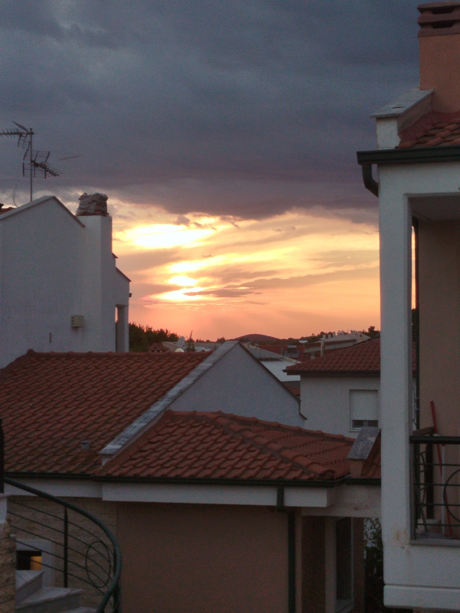 This is a vertical, medium-shot, eye-level photograph of a sunset over a residential area. The sky is a gradient of dark gray clouds at the top transitioning to bright yellow and orange hues closer to the horizon, indicating the setting sun. Several buildings with terracotta tile roofs are visible, with the closest one in the foreground on the left being a white, multi-story structure. A spiraling metal railing for an outdoor staircase is on the far left. The other buildings are of varying heights and architectural styles, also featuring tiled roofs. Some appear to be apartment buildings or houses. A television antenna is perched on top of a white building to the left. In the distance, a line of trees forms a dark silhouette against the vibrant sky. The overall impression is of a quiet evening in a coastal town, possibly during the warmer months. The location is identified as Sithonia Municipal Unit, Greece. There are no people visible in the image.