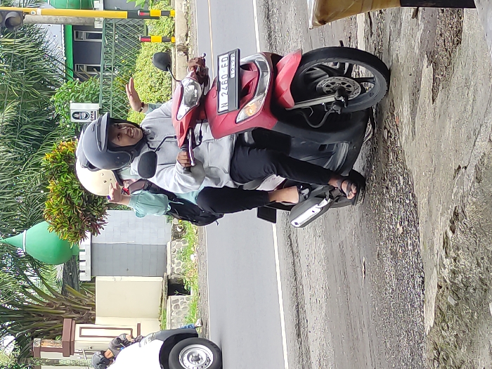This outdoor image, taken in Kepanjen, Indonesia, captures two individuals riding a red scooter on a road during what appears to be an overcast daytime. The rider, wearing a dark gray helmet, light jacket, and dark pants, smiles brightly at the camera while raising one hand in a wave. The passenger behind her, wearing a cream-colored helmet, light blue-green top, and dark pants, also smiles and gives a thumbs-up gesture. Both individuals appear to be female and exhibit a friendly, joyful demeanor.

The red scooter features a visible license plate displaying "N 2460 ED" and an expiry date of "12-23." The passenger is wearing black flip-flops. The background reveals a paved road with a white dividing line, flanked by lush green foliage, bushes, and palm trees. Concrete structures and parts of buildings are visible, including a prominent green dome structure, possibly belonging to a mosque. A traffic light pole is present in the upper left, and a white car is partially visible in the lower left corner. The soft, even lighting suggests an overcast sky, with no harsh shadows. A white sign with an unreadable black pictogram (possibly indicating a facility) is also visible in the left background.