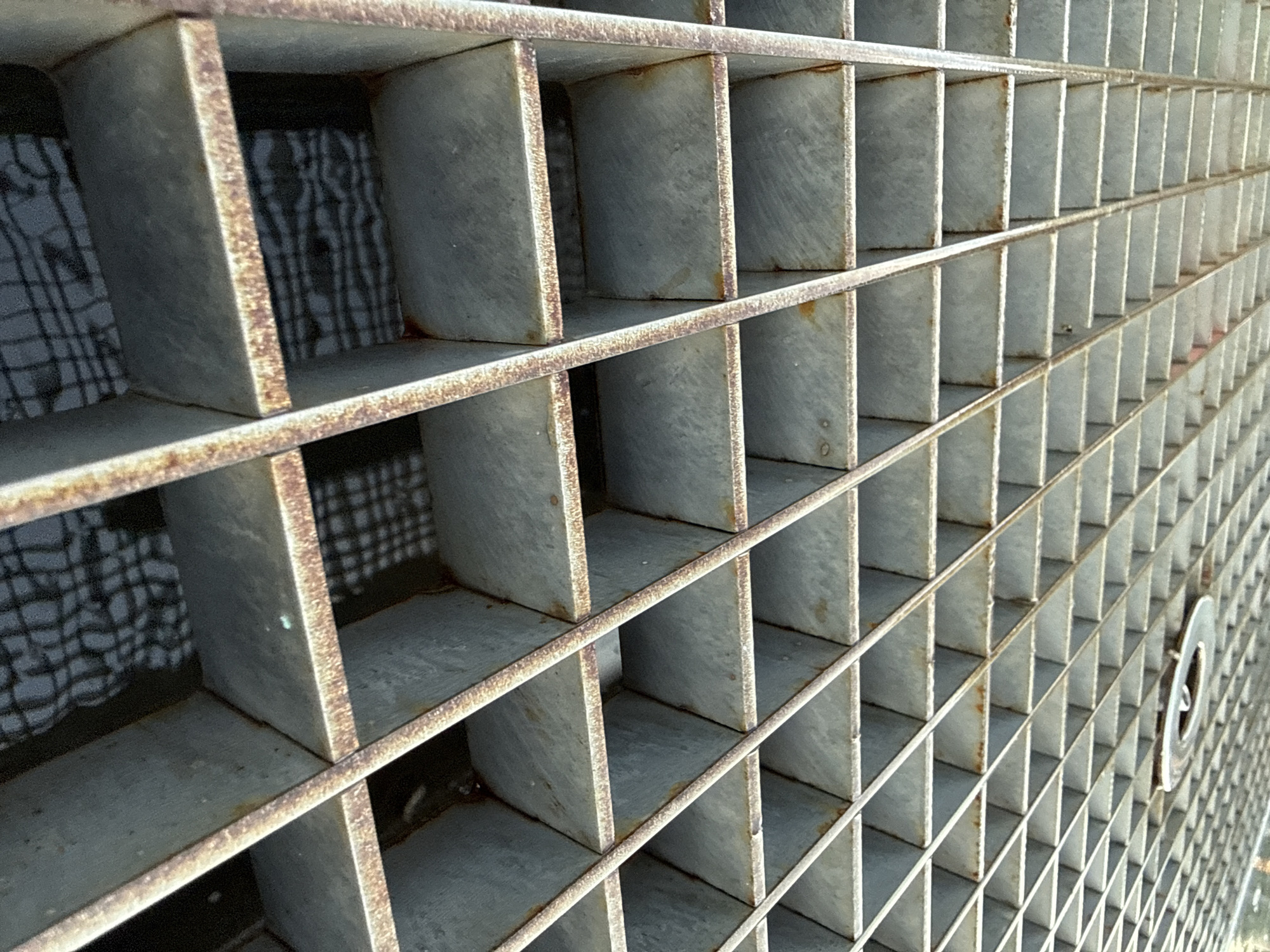 The image shows a close-up, angled view of a metal grate, likely a drainage cover, positioned outdoors. The grate is made of thick, weathered metal bars forming a grid pattern. Rust is visible along the edges and seams of the bars, indicating age and exposure to the elements. The perspective suggests the grate is on a horizontal surface, possibly a sidewalk or road. Through the openings of the grate, a darker surface is visible below, with a faint pattern that could be a reflection or another layer of grating. In the upper right portion of the image, a circular metal fixture, possibly part of the grate or a separate access point, is visible. The lighting is even, with no harsh shadows, suggesting an overcast day or diffused light. There are no people or immediate signs of activity in the frame. This grate is located in Piatykhatky, Ukraine.