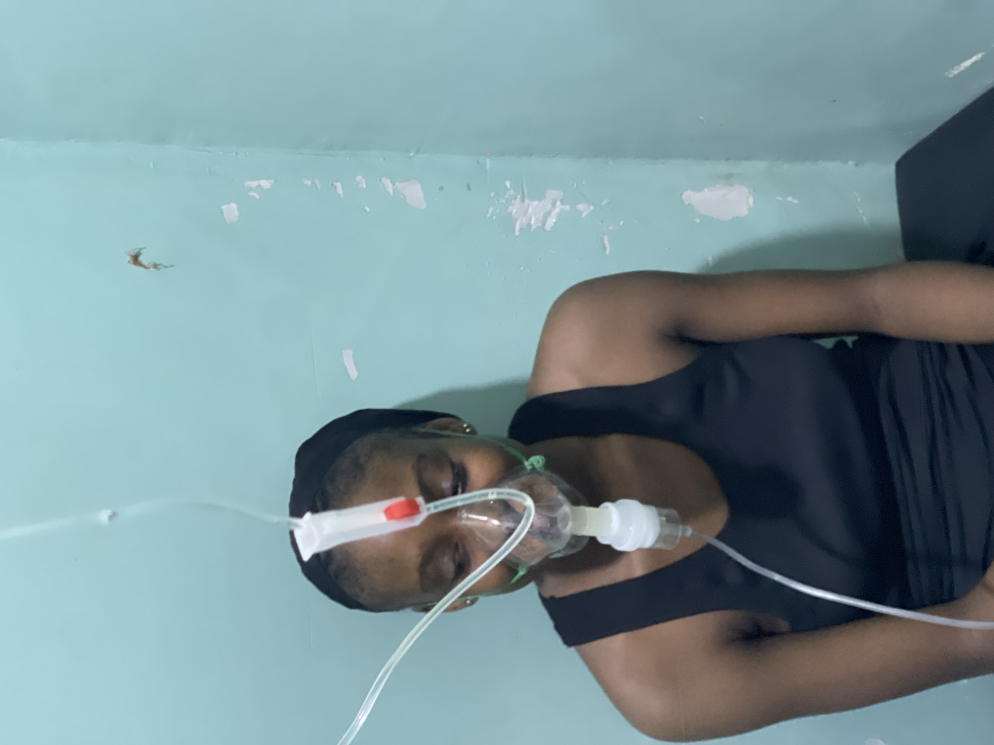 A young woman wearing a black tank top and a black headscarf is seated in front of a light blue, peeling wall in what appears to be a medical setting. She has a clear nasal cannula mask over her nose and mouth, connected by clear tubing to a nebulizer device. The nebulizer is attached to a white tube that disappears out of the frame, presumably connected to a larger oxygen source. The woman's eyes are cast downwards, and she has a slight smile. The wall behind her has patches of peeling paint and what looks like a spiderweb in the upper right corner. The lighting is dim, suggesting an indoor environment in Abuja, Nigeria.