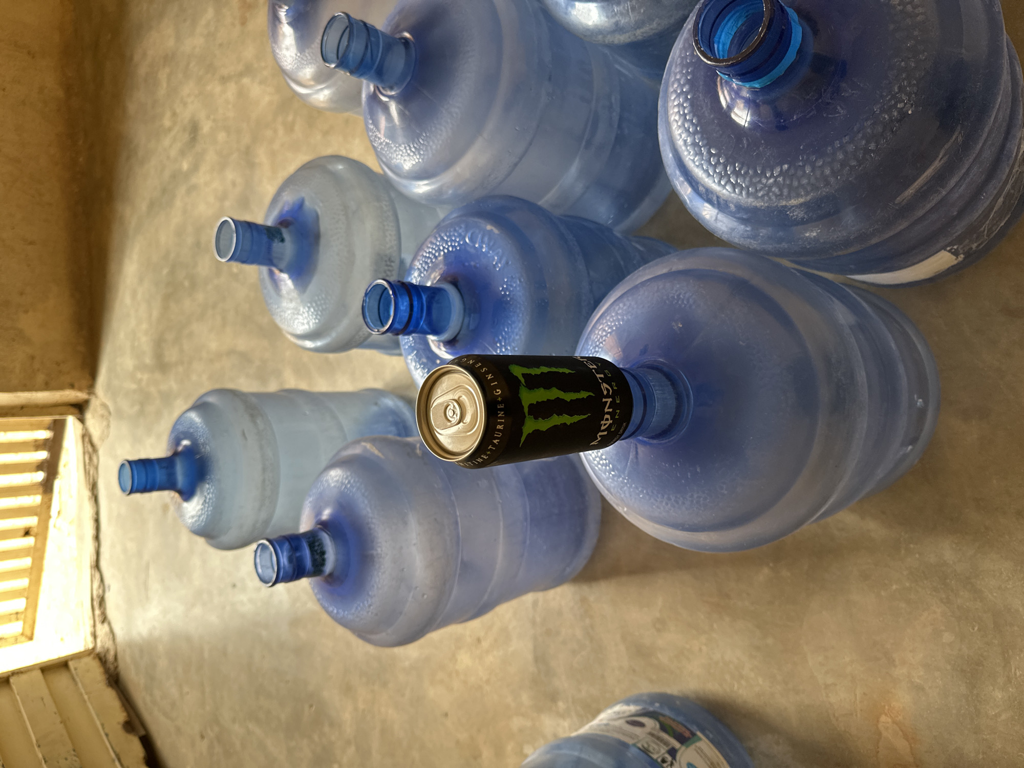 This overhead shot, taken in Jos, Nigeria, displays an assortment of large, blue plastic water bottles resting on a light brown, possibly concrete, floor. Most of the bottles appear to be empty or partially filled, evidenced by reflections and visible air bubbles. They are arranged somewhat haphazardly across the surface, suggesting a storage area or a collection point for refills.
A distinct black Monster Energy drink can is positioned upside down on the neck of one of the central water bottles. Clearly legible text on the can includes "MONSTER ENERGY" in its signature green, alongside "REHAB," "GINSENG," and "TAURINE" printed vertically. The can's silver top and pulled tab are visible.
The scene is brightly lit by natural daylight, casting highlights on the glossy surfaces of the bottles and the floor. To the left, a light-colored architectural element with horizontal slats, possibly a window frame or part of an interior wall, provides a structural boundary. No people are visible, and the overall impression is one of everyday utility or storage.