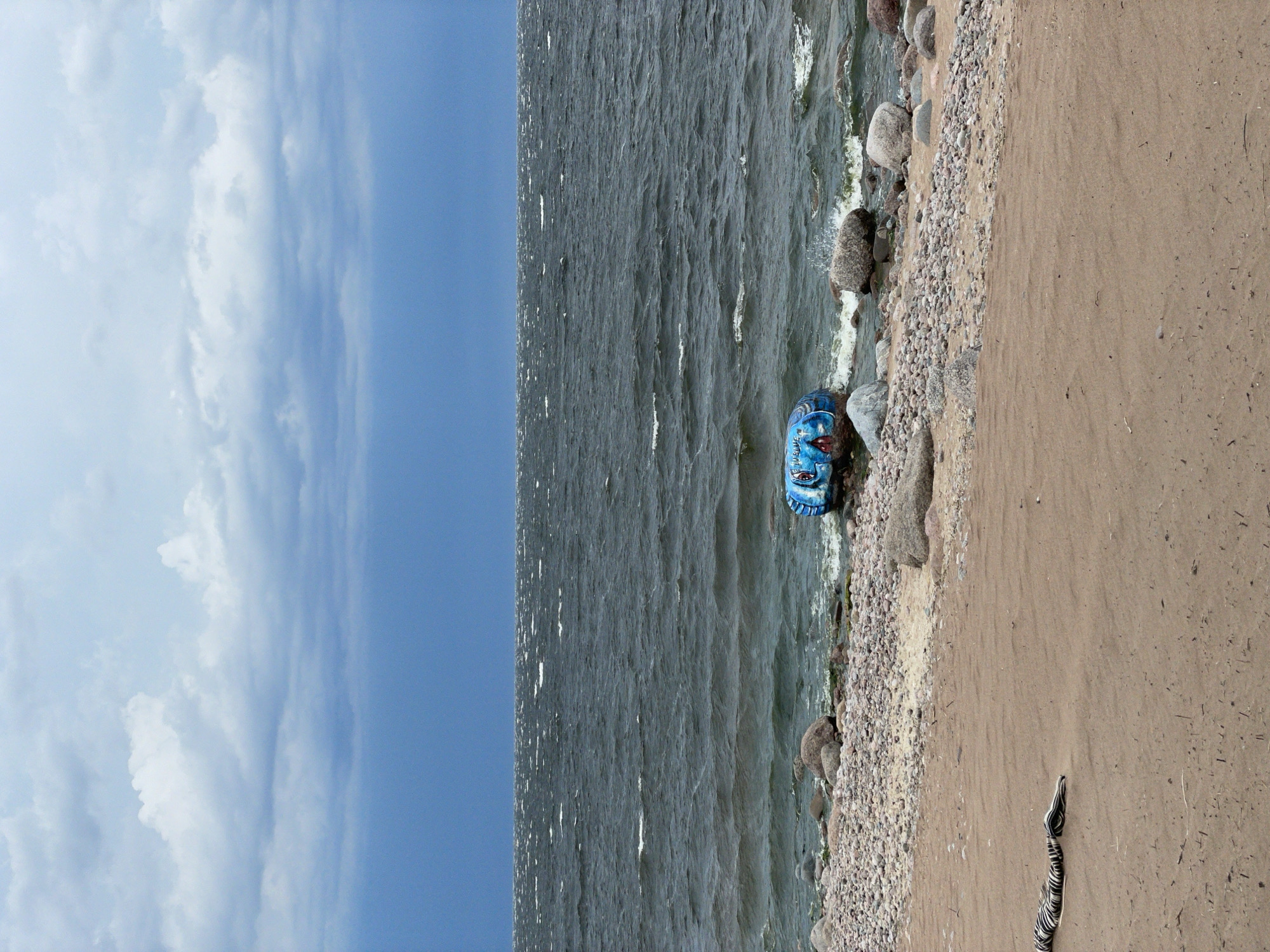 An inflatable tube with a painted fish face is partially submerged in choppy, dark water near the shore of a pebble and sand beach. The sky above is overcast with dramatic, dark clouds. The scene appears to be the shore of Luzhki, Russia, based on the context provided. A dark and white striped object, possibly a piece of fabric or clothing, lies on the sand in the foreground. Large, grey rocks are scattered along the shoreline. The water is restless, with whitecaps indicating strong winds.