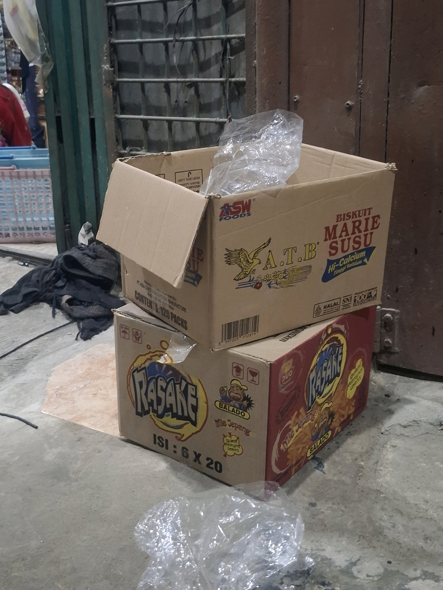 Two cardboard boxes are stacked on a concrete floor in an outdoor setting in the city of Medan, Indonesia. The top box is open and contains a crumpled plastic bag. It is labeled "BISKUIT MARIE SUSU Hi-Calcium". The bottom box is red and features branding for "RASAKE MIE JEPANG BALADO". The background shows a dark, barred metal structure and a brown wooden door. To the left, a portion of a blue plastic basket and some black fabric are visible. The lighting suggests it may be late afternoon or early evening.