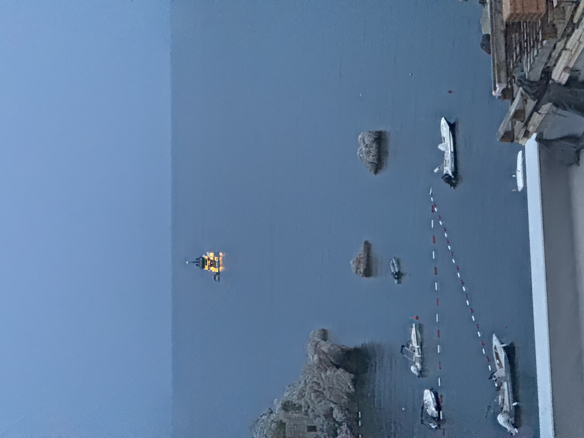 In Kamenovo, Montenegro, at dusk, a distant tugboat with illuminated decks is visible on the calm, dark blue sea. Closer to shore, a rocky outcrop juts out of the water, with the remains of a stone structure on its left. Several small boats are anchored in the bay, some resting on the water, others partially submerged. A rope barrier with red and white buoys delineates an area in the water. In the foreground, a modern building with exposed wooden beams and stacked bricks occupies the lower right corner of the frame.