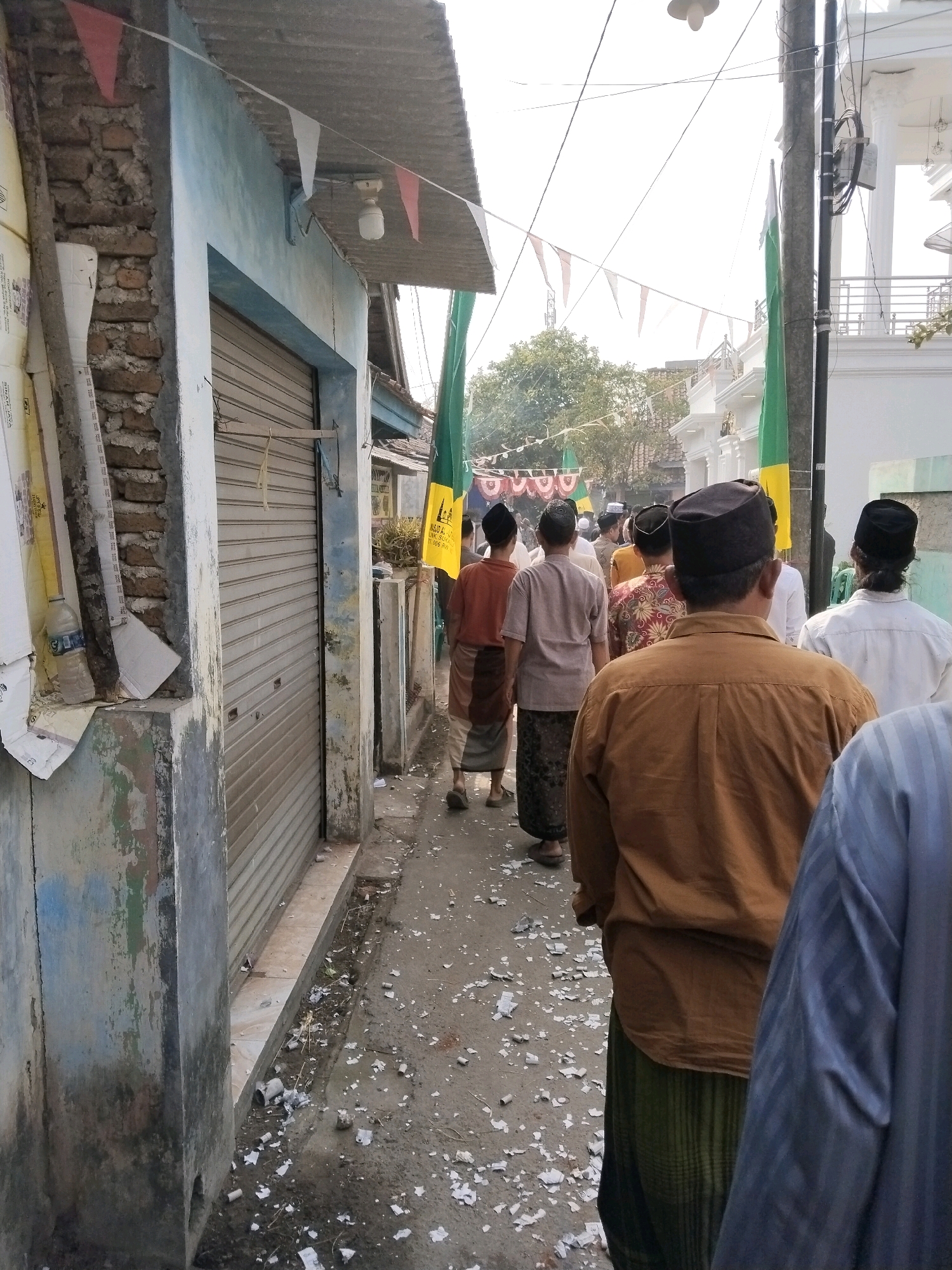 The image depicts a narrow, paved alleyway in the city of Serang, Indonesia. The alley is lined with buildings on both sides. On the left, a building features a light blue facade with a rolling metal door and exposed brickwork, showing signs of age and wear. Banners and flags are strung across the alley, including triangular red and white flags and green and yellow flags bearing text. A pole with electrical wires runs vertically along the right side of the alley. Scattered on the ground are pieces of white paper, suggesting a recent event or celebration.
A group of men, mostly wearing traditional Islamic attire, are walking down the alley away from the camera. They are diverse in age, with some wearing peci (traditional caps). One man in the foreground is wearing a brown long-sleeved shirt and a green sarong. Another man to his left wears a light brown shirt and a patterned sarong. Further down the alley, more men are visible, including one in a dark brown shirt and another in a light colored shirt. The overall atmosphere appears to be that of a procession or a gathering. The lighting suggests it is daytime, with the sun casting some shadows.