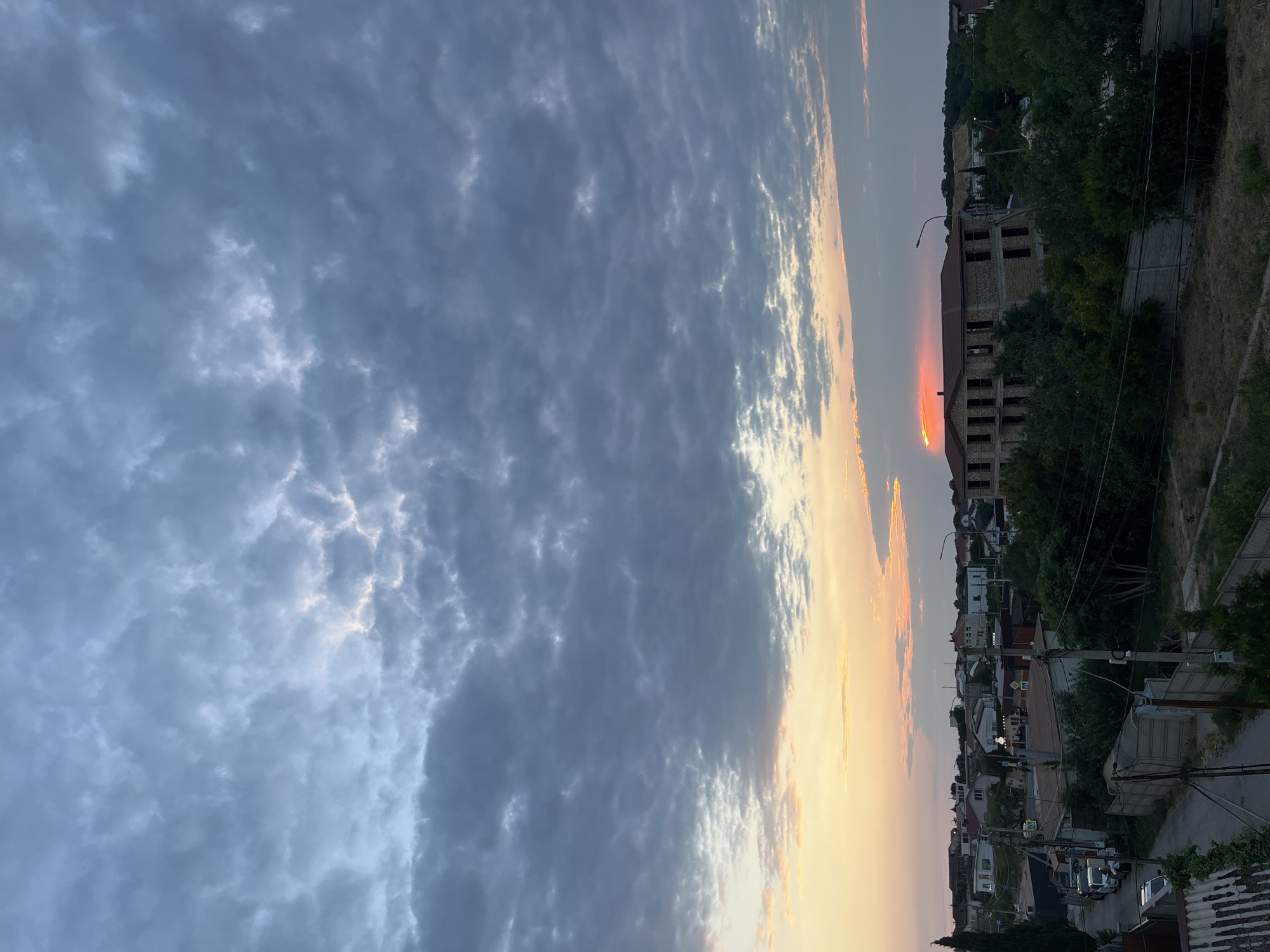 The image depicts a sunset over a residential area in Balaklavsky District, Ukraine. The sky is filled with dramatic, dark gray clouds, interspersed with streaks of orange and pink from the setting sun. A vibrant orange glow emanates from behind a large, unfinished brick building in the mid-ground. This building appears to be under construction, with exposed window frames and a dark roof.
The foreground shows a dirt road or pathway with a concrete fence on the right, and what appears to be a wooden fence or corrugated metal structure on the left. Several trees and dense green foliage surround the buildings, creating a lush, natural element within the urban landscape. In the distance, a line of houses with red and white roofs is visible, interspersed with trees and utility poles. Parked cars are lining the street on the left side of the image. The overall impression is one of a quiet, developing residential area experiencing a beautiful sunset.
