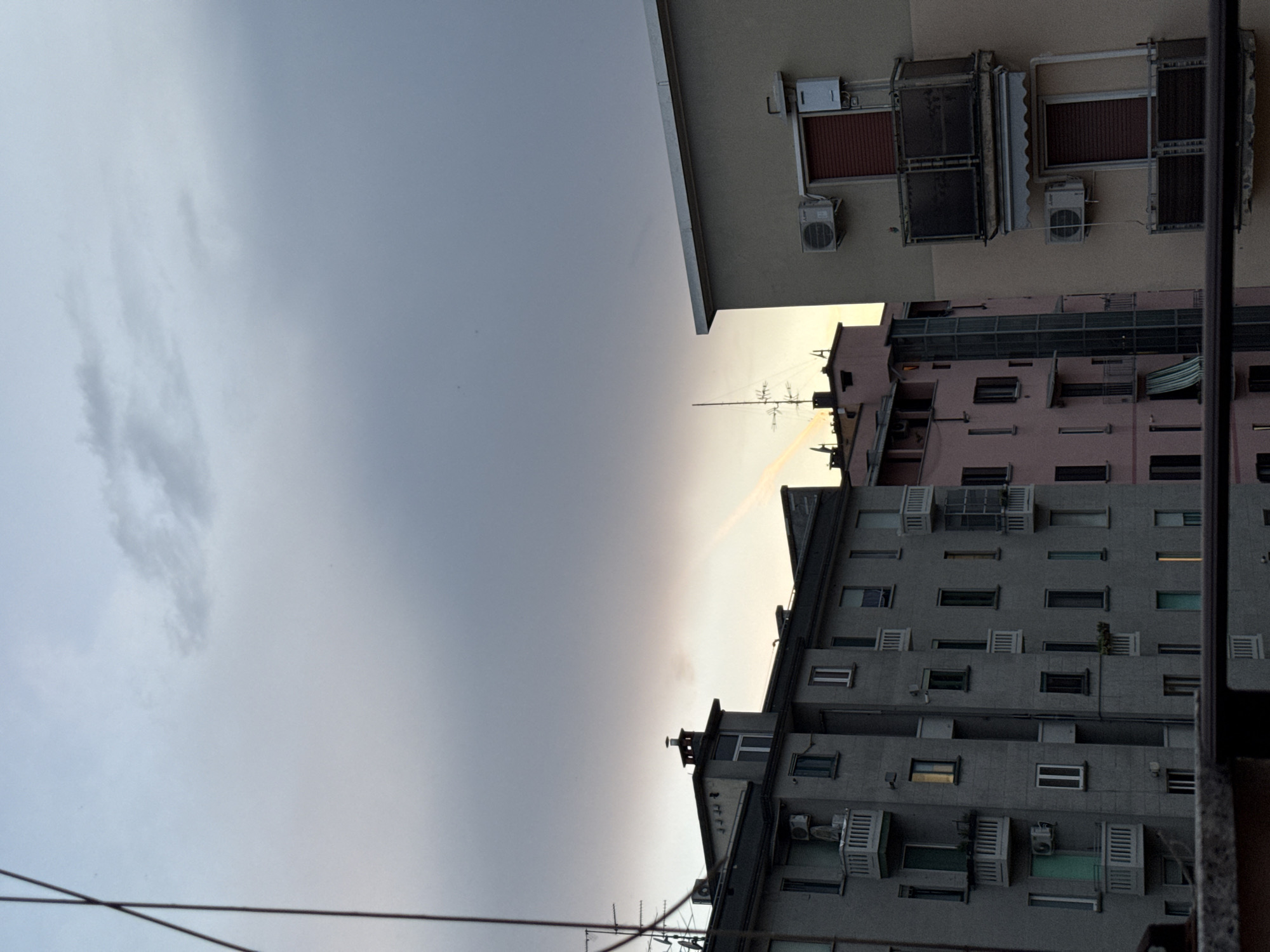The image shows a cloudy sky over apartment buildings in Milan, Italy, during twilight. A faint rainbow-like phenomenon is visible in the sky. The buildings are typical of urban residential architecture, with multiple floors, balconies, and windows. There are no people or animals visible in the scene. The foreground is occupied by a dark, out-of-focus railing, suggesting the photo was taken from a higher vantage point. The lighting indicates it is either dawn or dusk, with a mix of natural light and artificial lights from within some of the apartments. Antennas and air conditioning units are visible on the exteriors of the buildings.