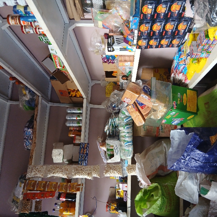 The image displays the interior of a retail general store in Moshi, Tanzania. The space is characterized by white metal-frame shelving units heavily laden with merchandise, extending along the visible walls.
On the upper left, multiple clear plastic bags of popcorn hang vertically alongside strips of orange-packaged snacks. Below these, various beverages, including Coca-Cola and Fanta, are arranged with stacks of white paper products and green-labeled aerosol cans.
The upper right shelves feature an assortment of packaged food items, including jars, cans, and cardboard boxes. An open cardboard box is also visible.
Mid-level and lower sections contain large green and white polypropylene sacks, appearing to hold bulk grains or pulses. One white sack has handwritten markings. A clear plastic container, filled with popcorn, rests on a green box labeled "BEN TEN." Stacked loaves of bread in clear plastic wrappers are positioned on top of the popcorn container.
The lower right shelving and counter display numerous small packaged goods, such as biscuits, candies, and 'POWER' branded boxes, likely containing batteries or small electronics. A crumpled blue plastic bag lies on the lower left.
The walls are painted a pale pink or purple hue, and the ceiling is white. No individuals are present, and no actions or interactions are depicted within the static scene.