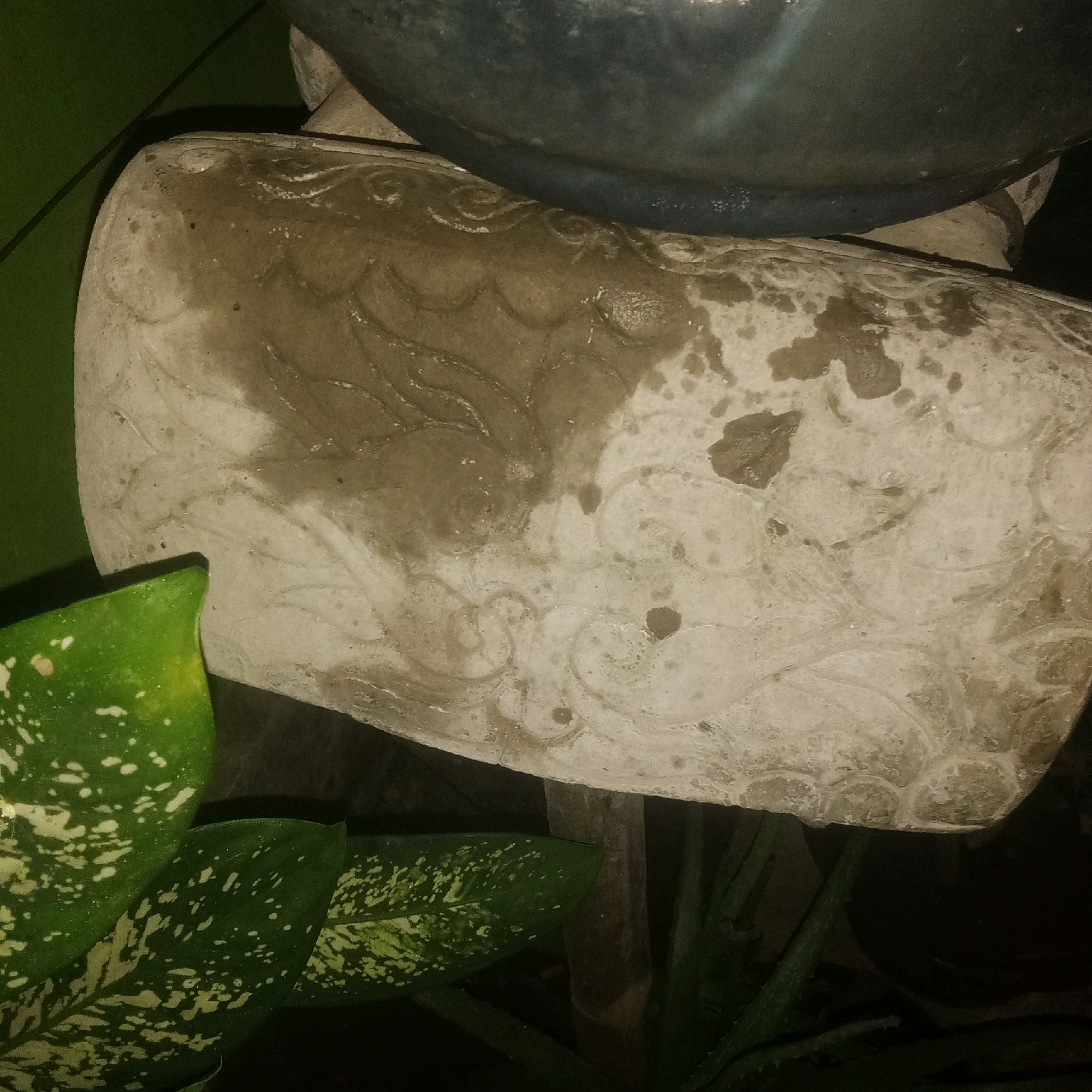 The image shows a close-up of a weathered, light-colored decorative concrete element, possibly part of a garden statue or planter, with embossed swirling patterns. The surface is mottled with darker, wet-looking patches and some lighter, dry areas. The top edge of this concrete element is visible, supporting a dark, possibly ceramic or metal object, likely a pot or bowl, which is partially out of frame. In the lower left corner, several green leaves with white speckles are visible, indicating the presence of a plant. The background is dark and out of focus, suggesting an outdoor setting at night or in a dimly lit area. There is no indication of the specific location within Karawang, Indonesia, other than what can be inferred from the general scene. There are no people or discernible actions depicted.