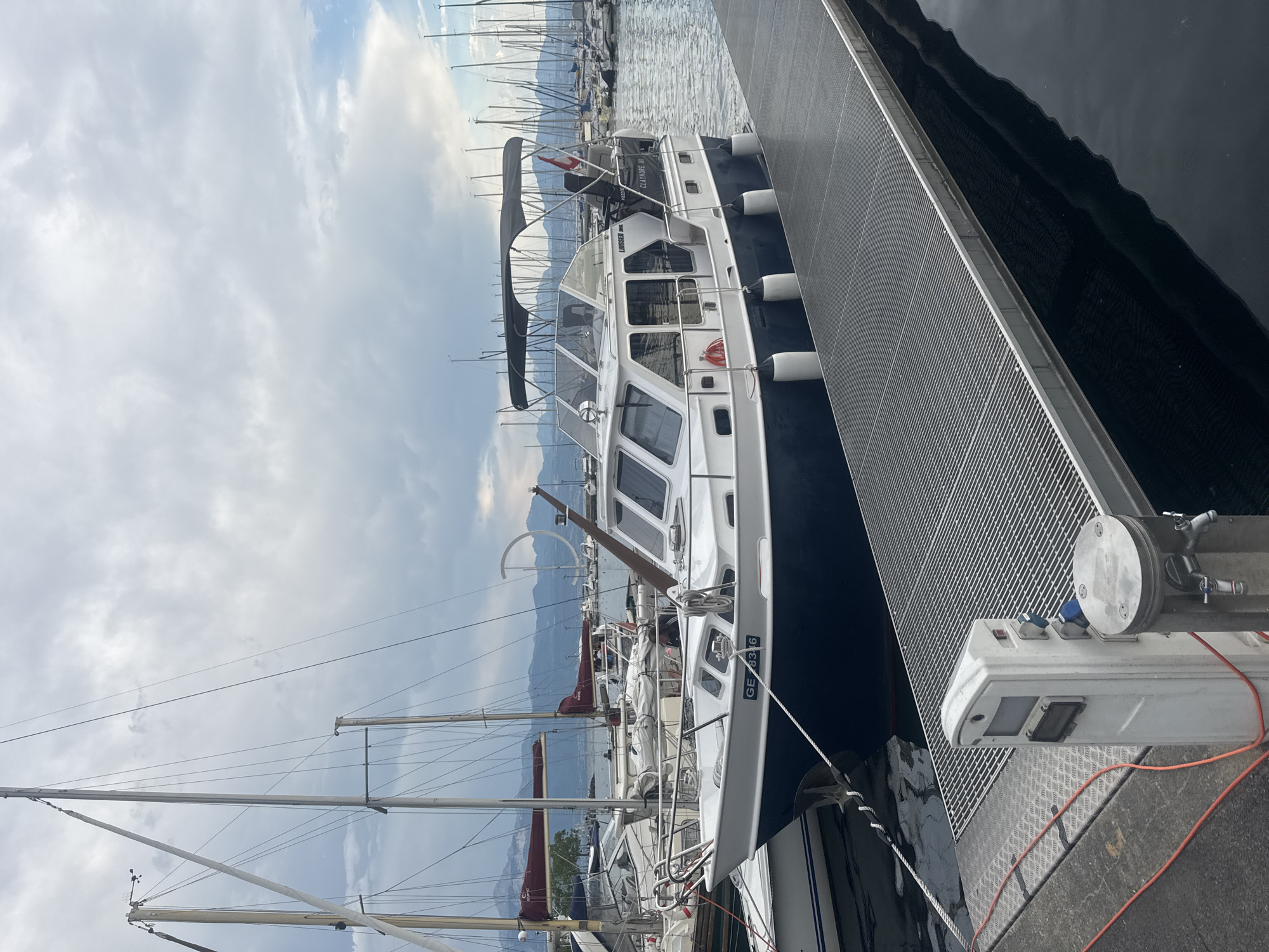 The image shows a motorboat docked at a marina in Lausanne, Switzerland. The boat is white with a dark blue hull and has several windows. It is docked alongside a metal gangway. In the background, there are numerous sailboats and yachts, as well as mountains and a cloudy sky. The marina appears to be located on Lake Geneva.