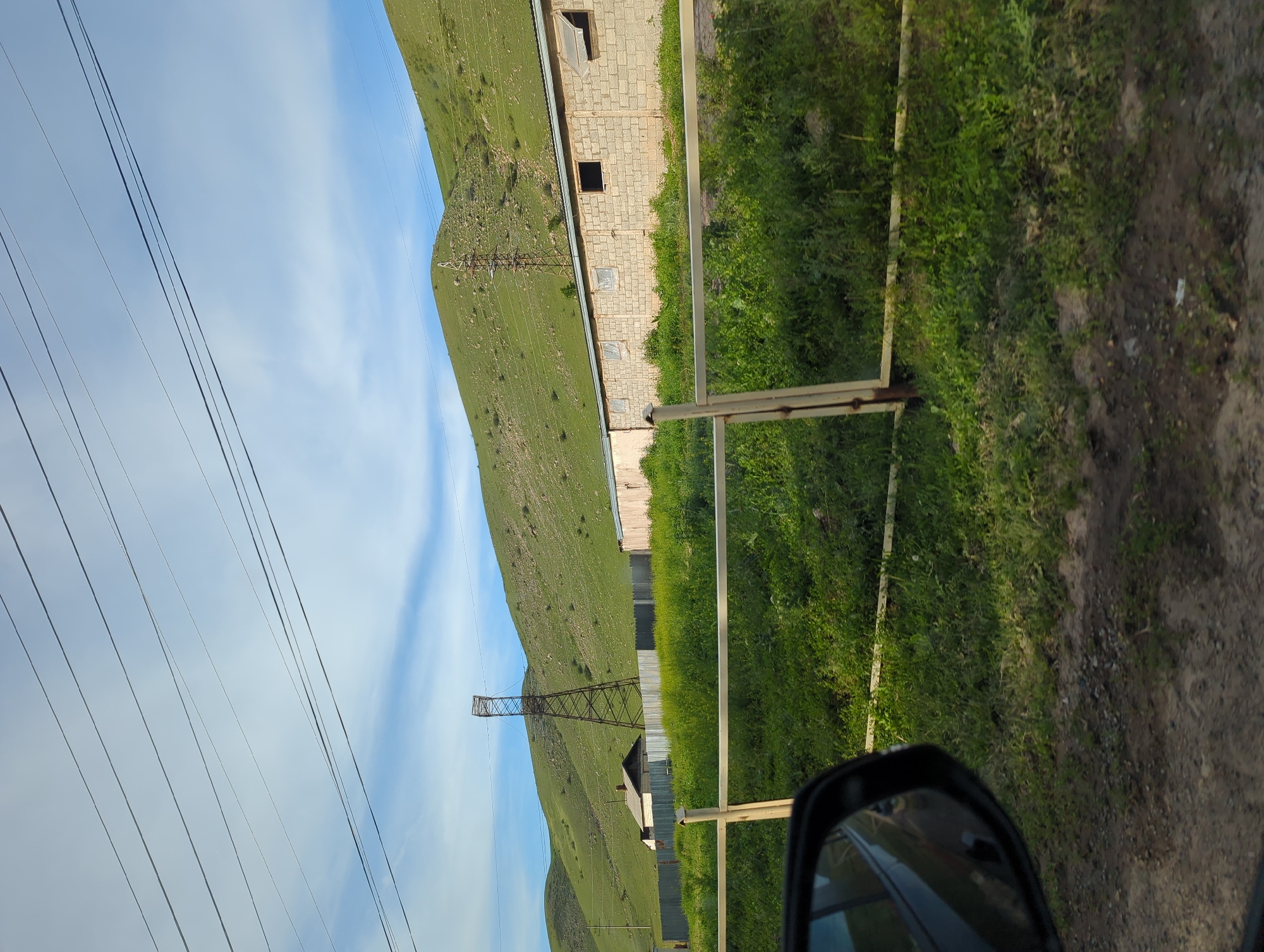 The image, apparently taken from a moving vehicle, shows a landscape in Koi-Tash, Kyrgyzstan. A lush, green hillside rises from the foreground on the left. An electrical transmission tower is visible against the sky and hill. Power lines stretch across the upper left corner.
Adjacent to the hill is a light-colored, brick-like building with visible windows. A grassy area extends in front of the building. In the foreground, a metal fence-like structure borders a dirt ground surface. A side-view mirror of the vehicle is visible in the lower right corner of the frame.