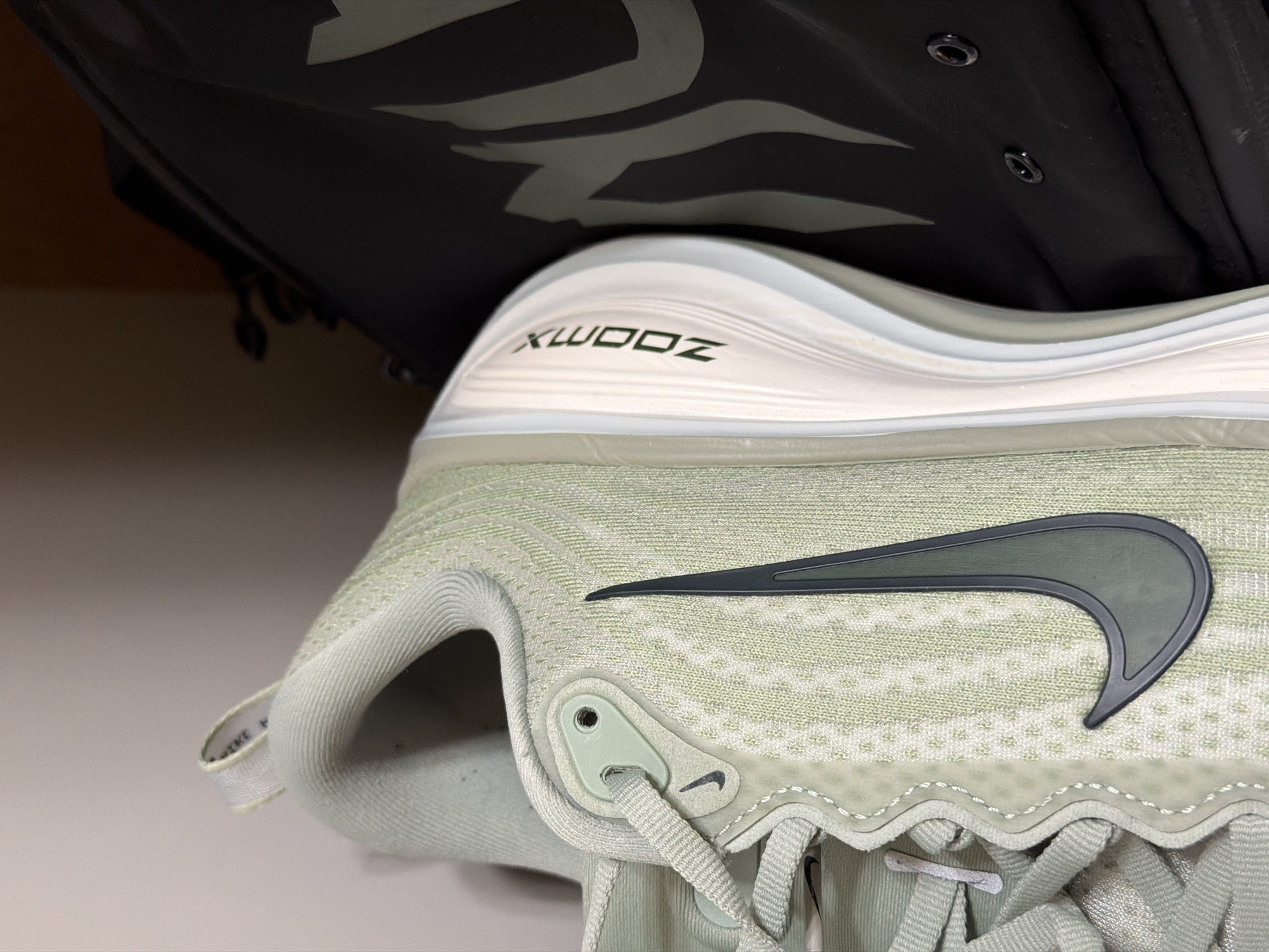 The image shows a close-up of a Nike shoe with a green upper and a black swoosh logo. The sole of the shoe has "ZOOM X" printed on it in black lettering. The shoe is partially obscured by a black garment with buttons visible near the top. The location in the city of Pleven, Bulgaria, is not ascertainable from the image.