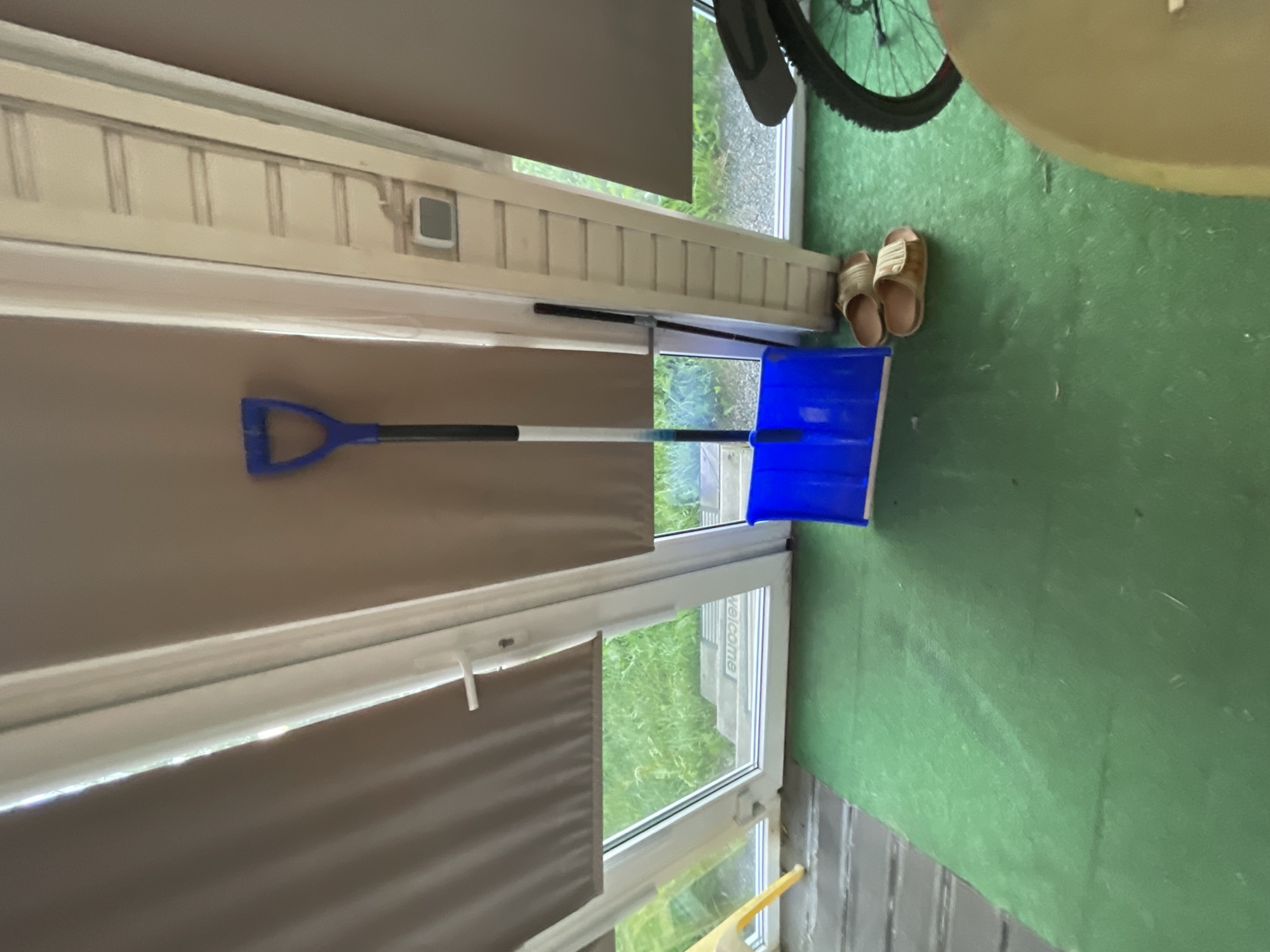 In what appears to be a sunroom or enclosed porch area in Bol'shoe Zaborod'e, Russia, a bright blue snow shovel leans against a wall near a set of sliding glass doors. The doors are covered by beige pull-down blinds, with a sliver of the outside greenery visible through the windows. On the green floor, a pair of tan flip-flops are placed next to the shovel. To the right, the rear wheel and part of the frame of a bicycle are visible, suggesting it is parked nearby. The overall impression is one of a functional, possibly utilitarian, space, perhaps preparing for or storing winter equipment.