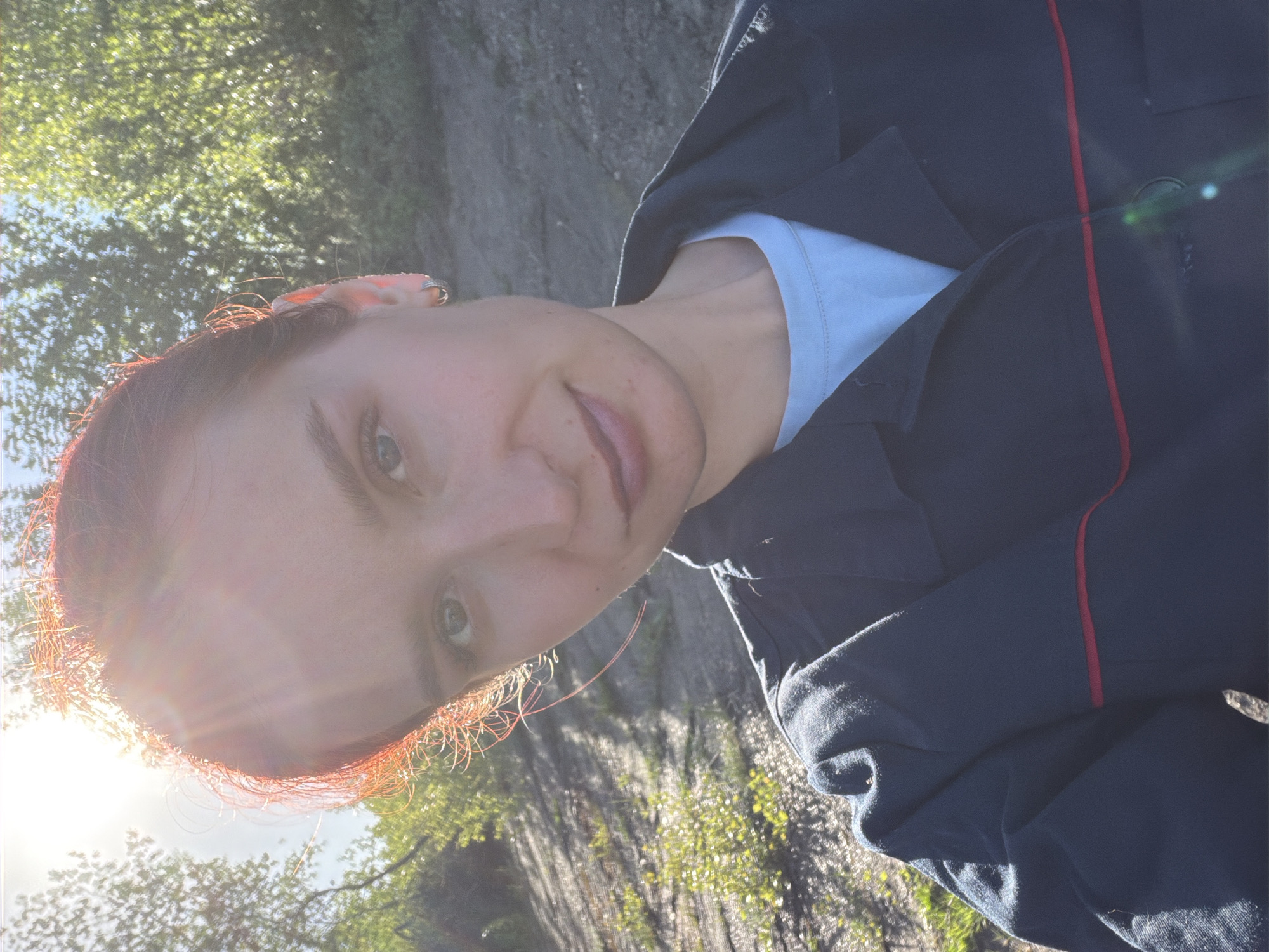 A person with red hair and blue eyes is shown in a selfie outdoors. The person is wearing a light blue t-shirt and a dark blue jacket with a thin red stripe. The background shows trees and sunlight. The location is potentially Chaikovsky, Russia, based on the prompt.