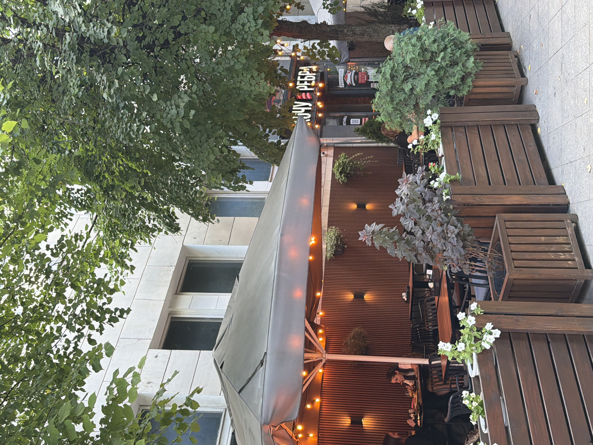 The image shows an outdoor seating area of a restaurant, likely in Zhytomyr, Ukraine, given the Cyrillic signage. The seating area is covered by a large gray umbrella with string lights illuminating the space. Wooden planters filled with greenery and flowers border the area. Inside the restaurant, visible through the entrance, is a neon sign with Cyrillic lettering. Several people are seated at tables, some eating and others engaged in conversation. The building behind the restaurant appears to be made of concrete and has large windows. The overall scene depicts a casual dining environment on a day with overcast or muted natural light.