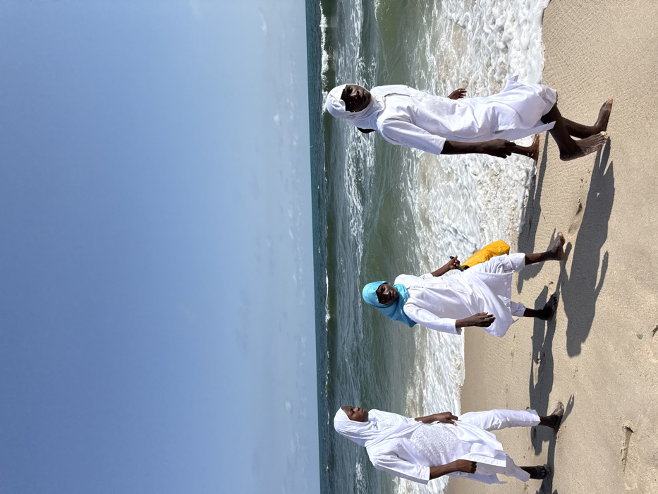 The scene is a sandy beach at Mopo Onijebu, Nigeria, under a clear, bright blue sky with faint clouds near the horizon. The ocean occupies the midground and background, exhibiting moderate waves breaking into white foam at the shoreline. The foreground consists of light brown sand, marked by footprints and long shadows cast by the individuals, indicating direct sunlight.
Four individuals are present. Three are prominently visible, walking barefoot on the wet sand near the water's edge, moving generally towards the right side of the frame. All three wear white long-sleeved tunics and white trousers.
The individual on the far left wears a white head covering and is seen in profile, looking towards the right. Their left foot is forward.
The middle individual wears a bright blue head covering, holds a yellow object in their left hand, and is looking towards the camera. Their right foot is forward.
The individual on the right wears a white head covering, which appears slightly disheveled around the forehead, and is looking towards the camera. Their left foot is forward.
A fourth individual is partially visible behind the rightmost person, with only a dark leg and foot discernible. They are also barefoot.
All visible individuals are in motion, traversing the interface between the beach and the ocean waves.