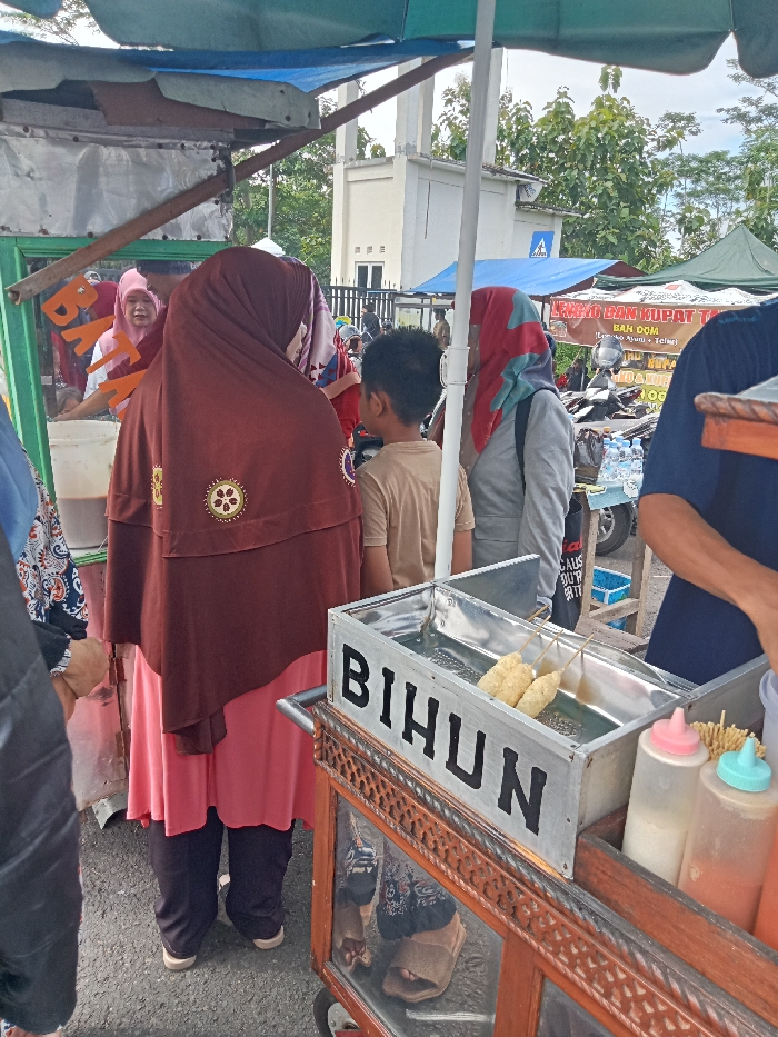 The image captures a vibrant street food scene in Singaparna, Indonesia, during the daytime. Several people are gathered around various food stalls.
In the foreground, a prominent wooden and metal food cart features a metal container with skewered items, likely fried snacks such as telur gulung (fried egg rolls), submerged in liquid or oil. The word "BIHUN" is clearly visible in large black letters on the side of this container, suggesting bihun (rice vermicelli) related items are sold, or that it's a general food cart. Next to it are two squeeze bottles with pink and light blue caps, presumably holding sauces, and a container of small fried stick snacks.
A woman in a brown hijab with circular embroidered patterns and a pink dress stands facing away from the viewer, looking towards a vendor on the left. Beside her, a young boy in a light brown t-shirt also stands with his back to the camera, observing the scene. Further to the right, another woman in a grey jacket and a red and blue patterned hijab is partially visible, carrying a black bag with "CALM UP FOR THE BATTLE" printed on it. On the far left, a vendor in a pink hijab is visible behind a stall with a white bucket. A green sign partially shows the word "BATAGOR," indicating another type of food available.
In the background, a white building with a distinctive structure, possibly a mosque minaret or a tower, stands under an overcast sky. Lush green trees frame the background. Another stall with a blue tarpaulin roof displays a sign with partial text, including "LANGSAT DAN KUPAT TA..." and "BAH DOM," indicating other traditional Indonesian foods or fruits are sold. A motorcycle and bottles of water are also visible on this stall. The overall atmosphere suggests a casual, bustling market or roadside vending area.