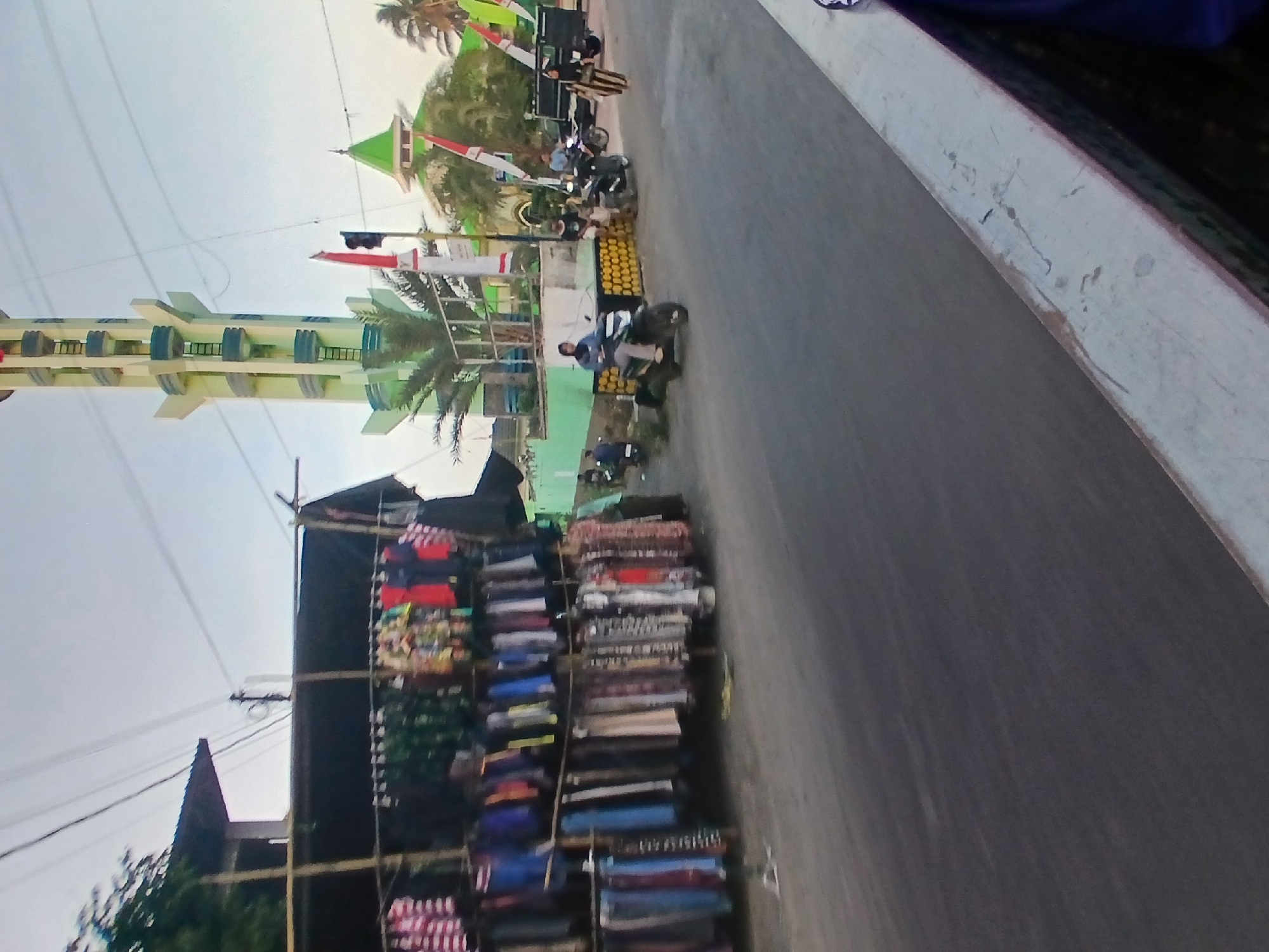 The image depicts a street scene in Wonosari, Indonesia. On the left side of the road, a market stall displays a variety of clothing items hanging from bamboo poles. The clothing includes shirts, dresses, and fabrics in various colors and patterns. Behind the stall, a building with a dark roof is visible, partially obscured by trees.
In the background, a tall, pale green and white tower, resembling a minaret, stands prominently against a clear sky. To the right of the tower, a building with a green roof, possibly a mosque, is visible. Several Indonesian flags are mounted on poles along the street.
The road is paved and appears to be a public thoroughfare. Several motorcycles are present on the road. One person is riding a motorcycle in the foreground, wearing a blue shirt and a helmet. Another person is walking on the right side of the road, wearing a striped outfit and a headscarf. A few other individuals are seen on motorcycles in the middle ground, and some are gathered near the buildings. The overall environment suggests a moderately busy street with a mix of commercial activity and local infrastructure. The lighting indicates it is daytime.
