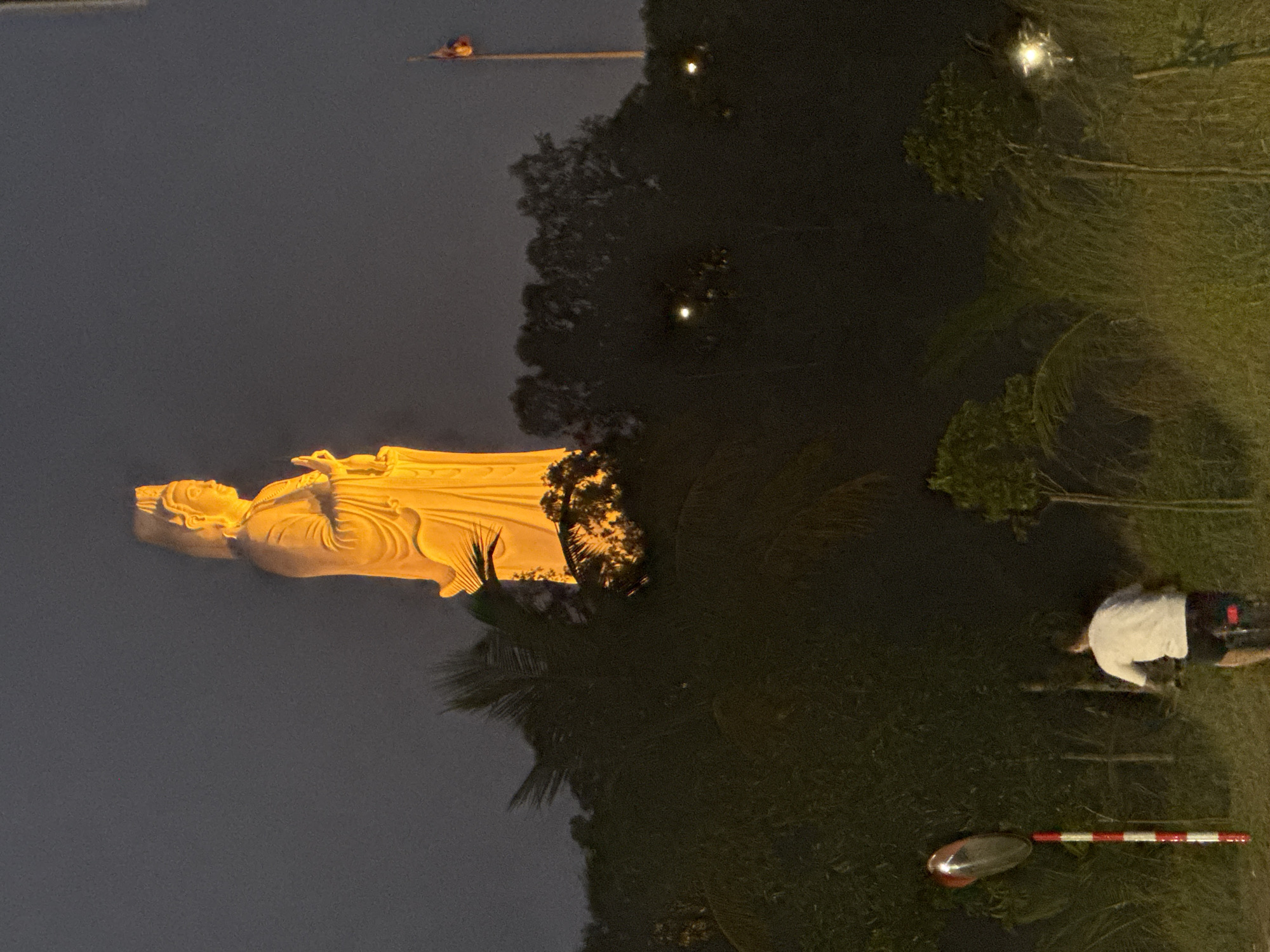 The image shows a nighttime view of the Lady Buddha statue at the Linh Ung Pagoda in Đà Nẵng, Vietnam. The statue is illuminated in a warm yellow light and stands on the shore of a body of water. To the right of the statue is a dark, dense area of trees and vegetation. In the lower right corner, a person wearing a white shirt and dark shorts is near some type of barrier, post, or sign. A red-and-white striped object lies on the ground nearby, and some sort of reflective object is further down. Further up and slightly to the left of the statue, a distant figure is seen on a small platform.