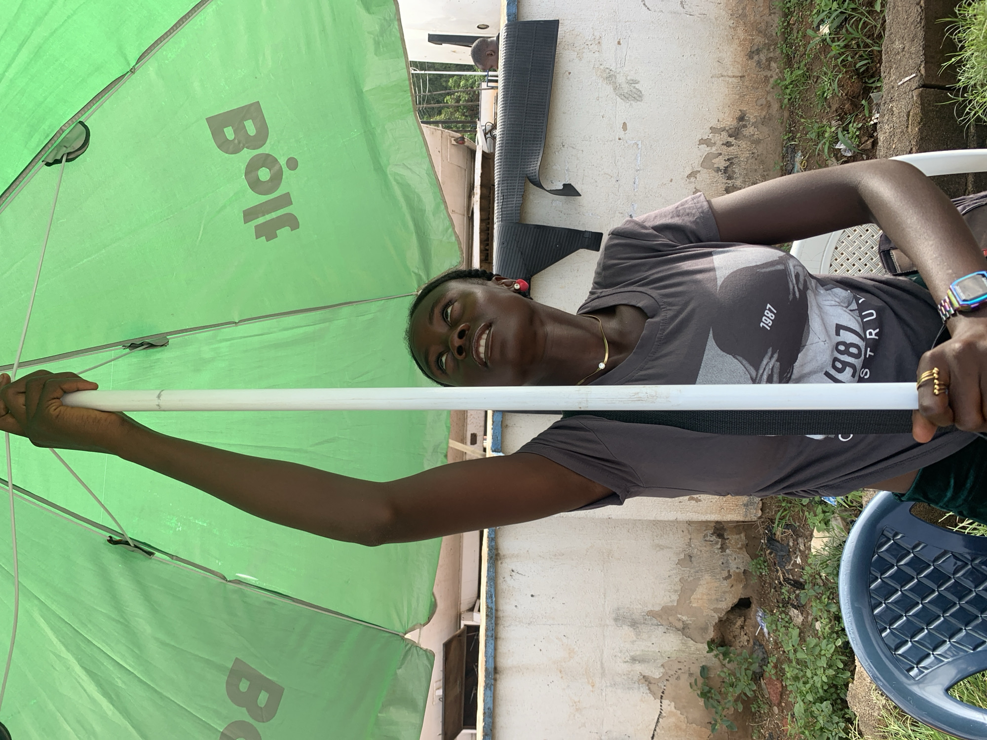 A young Black woman is captured outdoors, seated under a large, green umbrella with the "Bo" logo visible. She is holding the umbrella's central pole with her right hand, which is extended upwards. She is wearing a dark grey t-shirt with a graphic print, a gold necklace, and a red heart-shaped earring. Her left hand rests on her lap, adorned with gold rings. A wristwatch with a colorful band is visible on her left wrist.
She is seated on a blue plastic chair, with a portion of a white plastic chair visible on her right. The background consists of a textured, light-colored wall, with some overgrown vegetation and debris at its base. In the distance, a small section of what appears to be a building or structure is visible. The overall impression is an outdoor setting, possibly a market or roadside stall, in a part of the city of Jos, Nigeria.