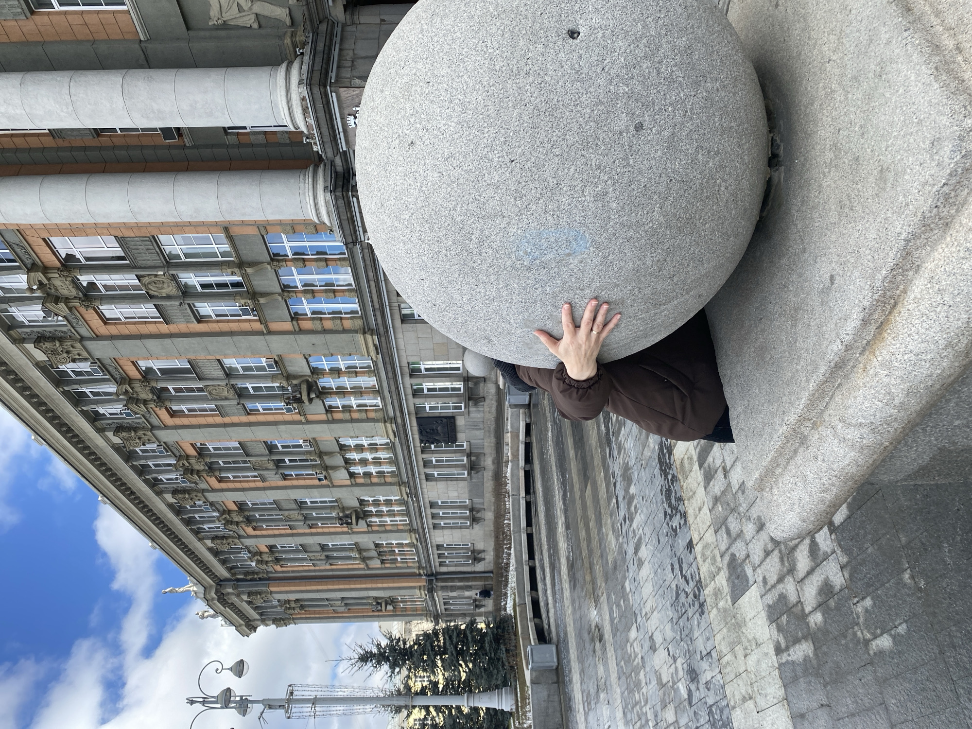 The photograph captures a scene in Yekaterinburg, Russia, featuring a large, spherical stone sculpture set against the backdrop of a prominent building. A person stands partially concealed beneath the sphere, their hand resting on its surface. The building behind features a classical architectural style, with multiple rows of windows, columns, and decorative elements. To the left, the sky is visible with a mix of blue and white clouds. Below the building, there's a tree, a lamp post, and a water feature. The ground is paved with stone slabs.