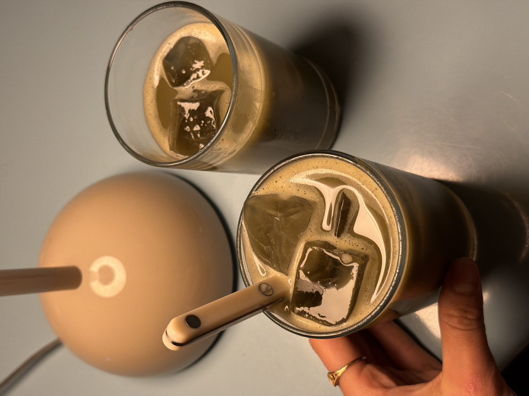 Two cylindrical glasses filled with a frothy, iced beverage, possibly coffee or matcha, are presented on a light blue surface alongside a beige-colored, round lamp. One glass is being held by a person's hand adorned with a gold ring. A small, beige-handled utensil is inserted into the beverage in this glass. The setting is indoors, illuminated by the lamp, which casts distinct shadows on the surface. There are no visible indicators suggesting the location is specifically in Krasnoyarsk, Russia.