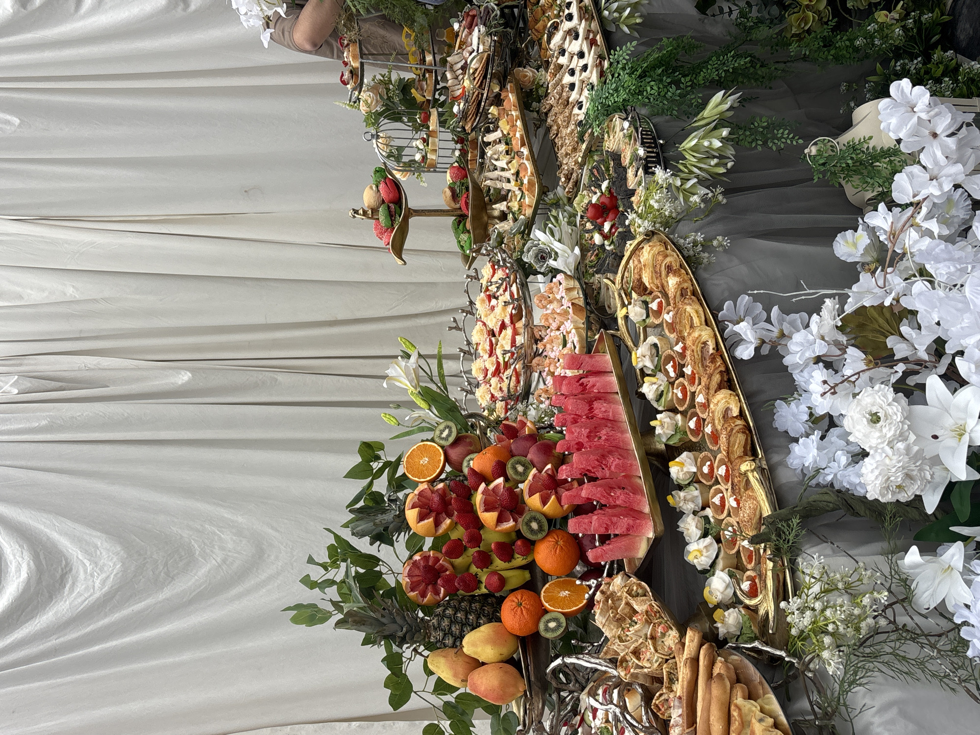 A high-angle shot displays a lavish spread of food decoratively arranged on tiered platters and interspersed with floral arrangements. The image showcases a variety of fruits including watermelon slices, oranges, kiwis, and bananas. The fruit is interspersed with an assortment of savory items such as rolls, breadsticks, and hors d'oeuvres. The presentation is enhanced by the inclusion of white and green floral arrangements. The backdrop is a draped white fabric. There is no evidence to connect the image with Yerevan, Armenia.