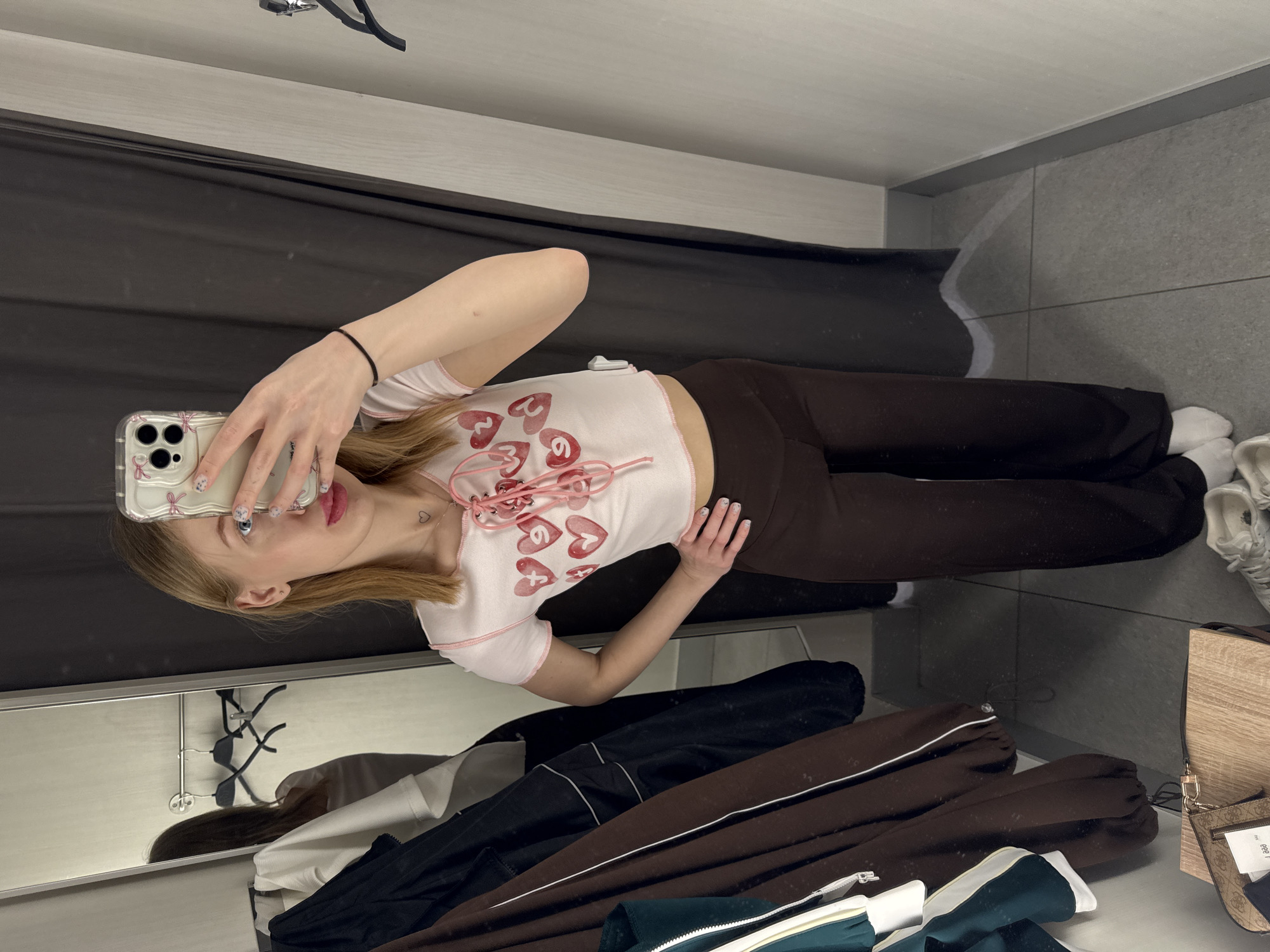 A fair-skinned female with blonde hair stands in an indoor fitting room in Ovoshchnoy, Russia. She is taking a mirror selfie, holding a smartphone with a clear, bow-decorated case in her raised right hand, partially obscuring her face. Her left hand is placed on her hip. She wears a light pink short-sleeved crop top with multiple red heart designs and pink lacing down the front, dark brown wide-leg trousers, white socks, and white sneakers. A small heart tattoo is visible on her lower neck. A black elastic band is on her left wrist. The room features light-colored walls, a dark grey tiled floor, and a full-length mirror reflecting the subject and two empty black clothing hangers on a rail. A dark grey privacy curtain is partially visible on the right. To the left, several items of clothing hang, including a dark jacket, a white shirt, a green and white striped garment, and a brown garment with a white stripe. A light-colored wooden surface in the bottom left corner holds a patterned "Guess" purse with a small white tag.