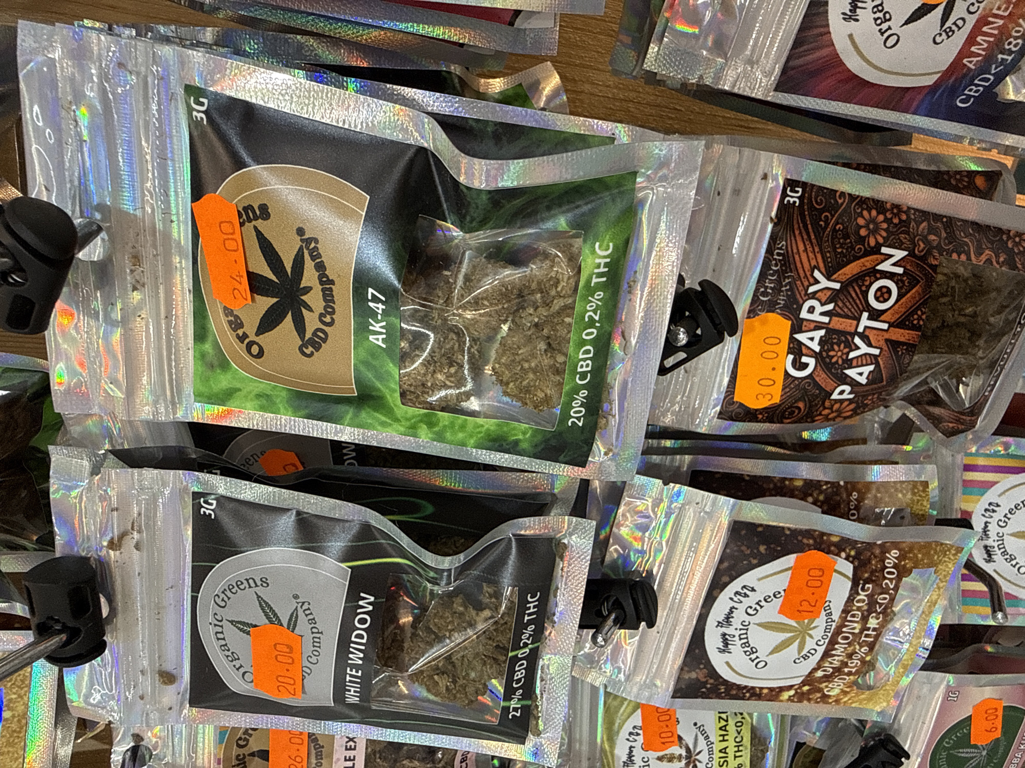 The image shows a retail display of packaged dried plant material, likely cannabis products, in what appears to be a shop. The products are packaged in small, resealable plastic bags with holographic or metallic finishes. Each bag features branding, product names such as "White Widow," "AK-47," and "Gary Payton," and nutritional information like CBD and THC percentages. Some bags also have orange price tags indicating monetary values such as "20.00," "24.00," "12.00," and "30.00." The products are hung from a display rack, with multiple rows visible. The background is blurry but suggests an indoor environment with wooden shelving or walls. There are no people visible in the image. The location is specified as Cales De Mallorca, Spain.