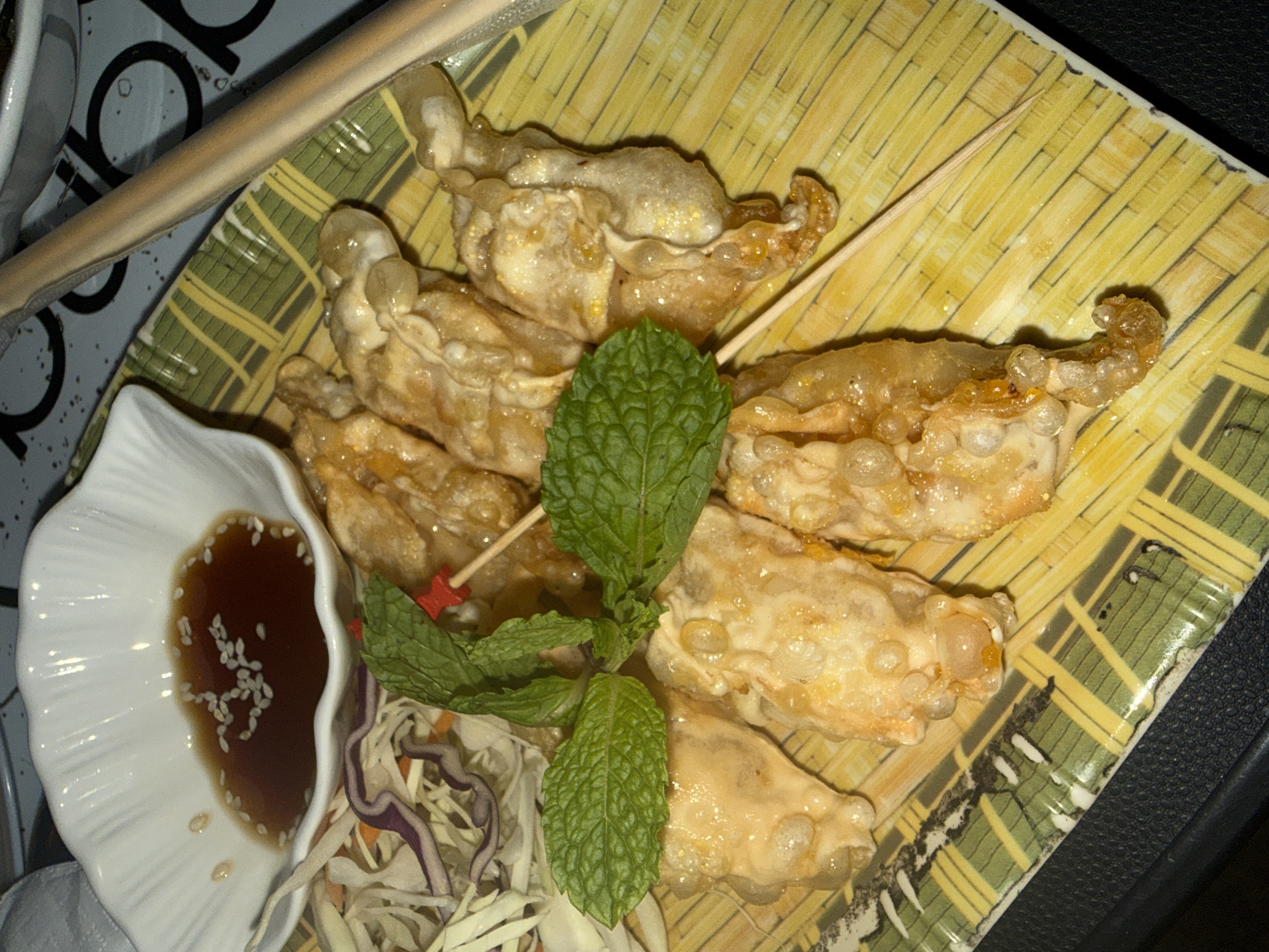 A plate of fried dumplings, a small bowl of dipping sauce with sesame seeds, and a side of shredded cabbage and purple cabbage are arranged on a yellow and green patterned plate. The dumplings are golden brown and appear to be coated in a glaze. They are garnished with fresh mint leaves and a red-tipped toothpick. Two chopsticks rest on the side of the plate. The scene is set on a dark, textured surface. The image does not provide any information about its location in Sakhu, Thailand.