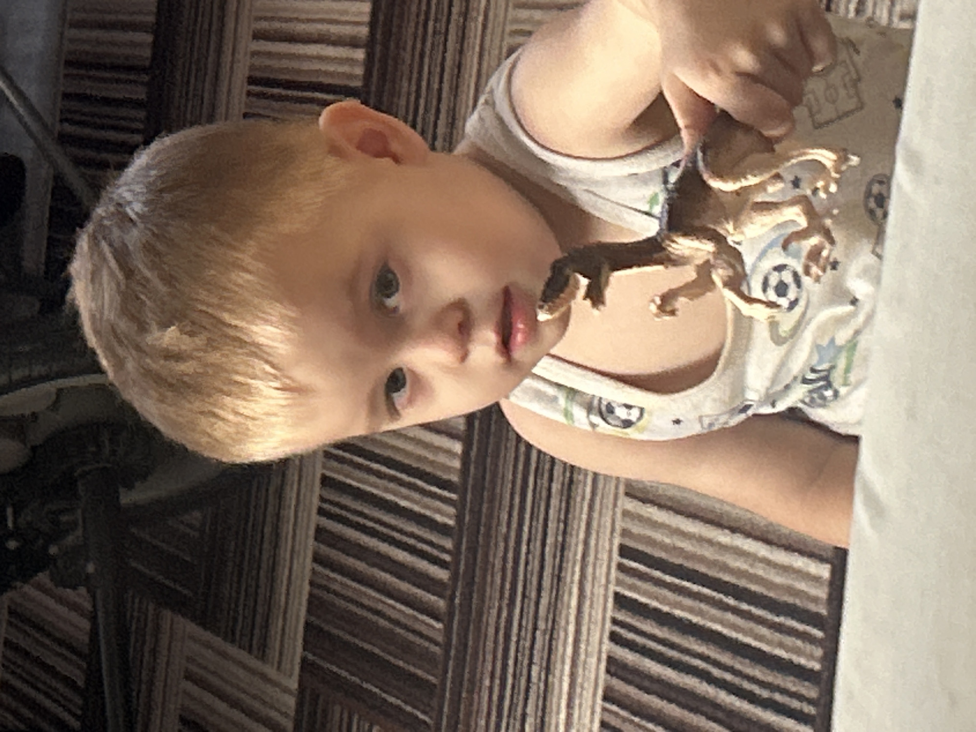 A young child, approximately two to three years old, with light blonde hair and light-colored eyes, is depicted in a medium close-up shot indoors in the city of Ольховка, Russia. The child wears a light-colored sleeveless top patterned with soccer balls and stars. The child's mouth is slightly ajar, and they are looking slightly to the left of the camera. A light brown, bipedal dinosaur toy is held in front of the child's chest by a hand, which has a ring on its fourth finger. The background consists of a patterned surface, likely a floor covering, featuring horizontal and diagonal stripes in shades of brown, beige, and dark grey. The lower foreground shows an out-of-focus light-colored surface.