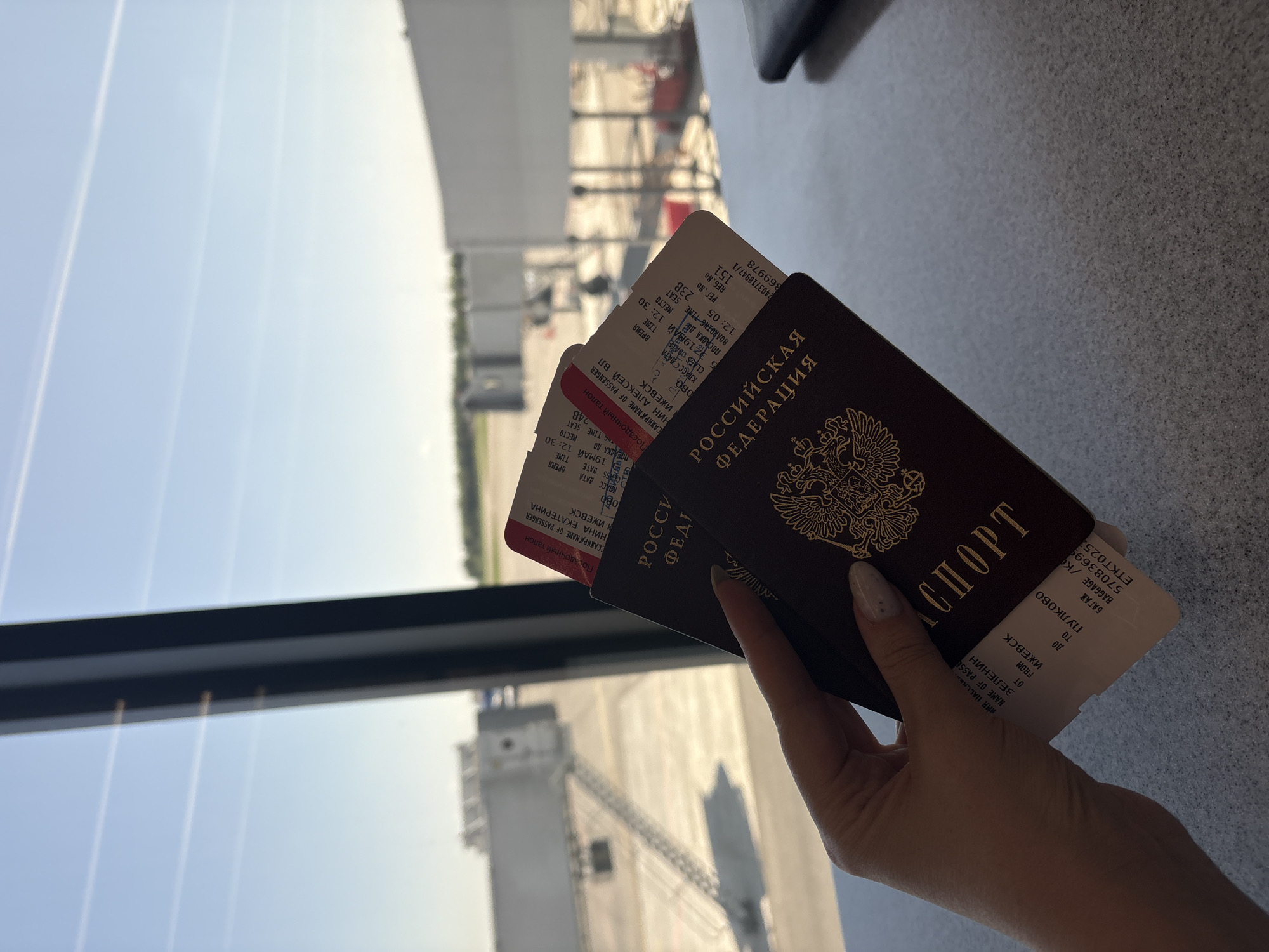 A hand holds two Russian passports and two boarding passes in front of a window at an airport. The passports are dark red with gold lettering. The boarding passes have Russian text. The airport window reveals a view of the airfield outside. The boarding passes indicate flights from Izhevsk to Pulkovo Airport. The context suggests the location might be Novoe Martyanovo, Russia, near Izhevsk.