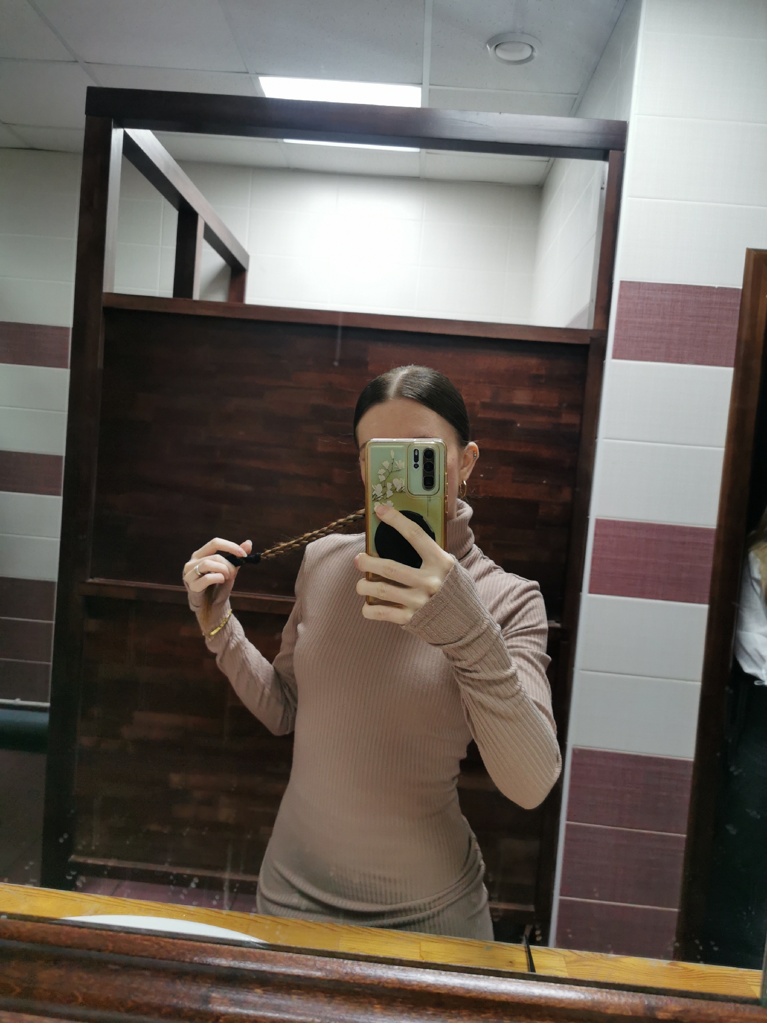 A woman is captured in a mirror selfie, standing in what appears to be an indoor, public or semi-public facility, possibly a restroom or changing room. She is positioned in front of a mirror, which reflects her image and the surroundings.
The woman has light brown hair, pulled back into a low ponytail or bun. She is wearing a light brown or beige ribbed turtleneck top. Her left hand is holding her phone, which has a light green case with a floral design, and she is taking the picture. Her right hand is visible, holding a braid of her own hair. On her left hand, she wears a silver-colored ring on her ring finger, and a gold-colored bracelet is visible on her right wrist. She also has small hoop earrings.
The setting behind her, reflected in the mirror, features wooden partitions, possibly defining stalls or changing areas. The walls are partly tiled with white square tiles, and below a certain height, there are horizontal stripes of a reddish-brown and white tiled pattern. The ceiling, visible in the reflection, consists of white dropped ceiling tiles with recessed fluorescent lighting fixtures and a circular vent. The mirror itself has a thick, dark wooden frame. There is no visible text in the image.