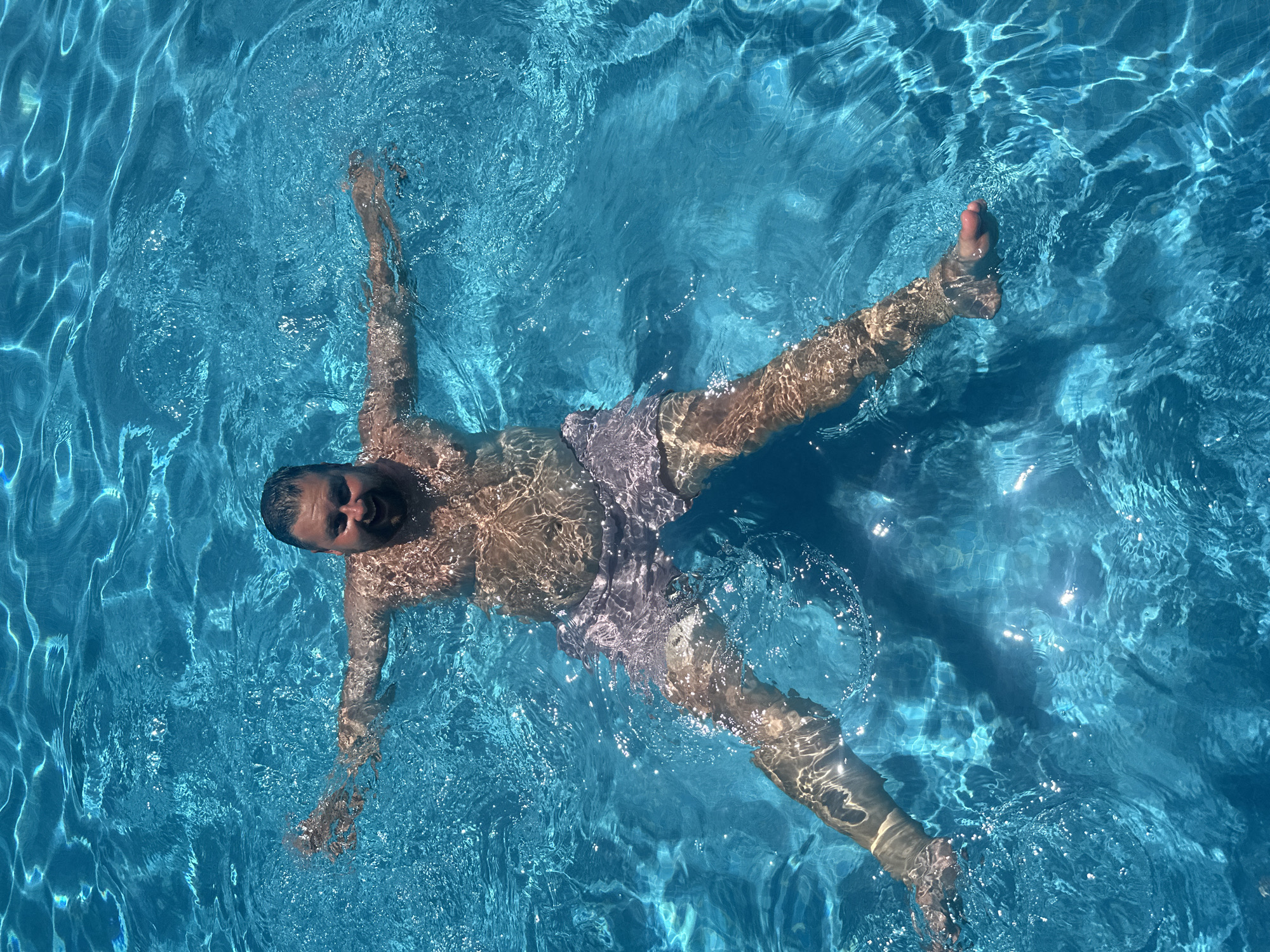 A man is floating on his back in a swimming pool in Sant Feliu de Guíxols, Spain. The water is clear and blue, with sunlight creating reflections on the surface. The man is smiling and has his arms and legs spread wide, appearing relaxed. He is wearing light-colored swim trunks. The perspective is from directly above the pool, looking down on the man.