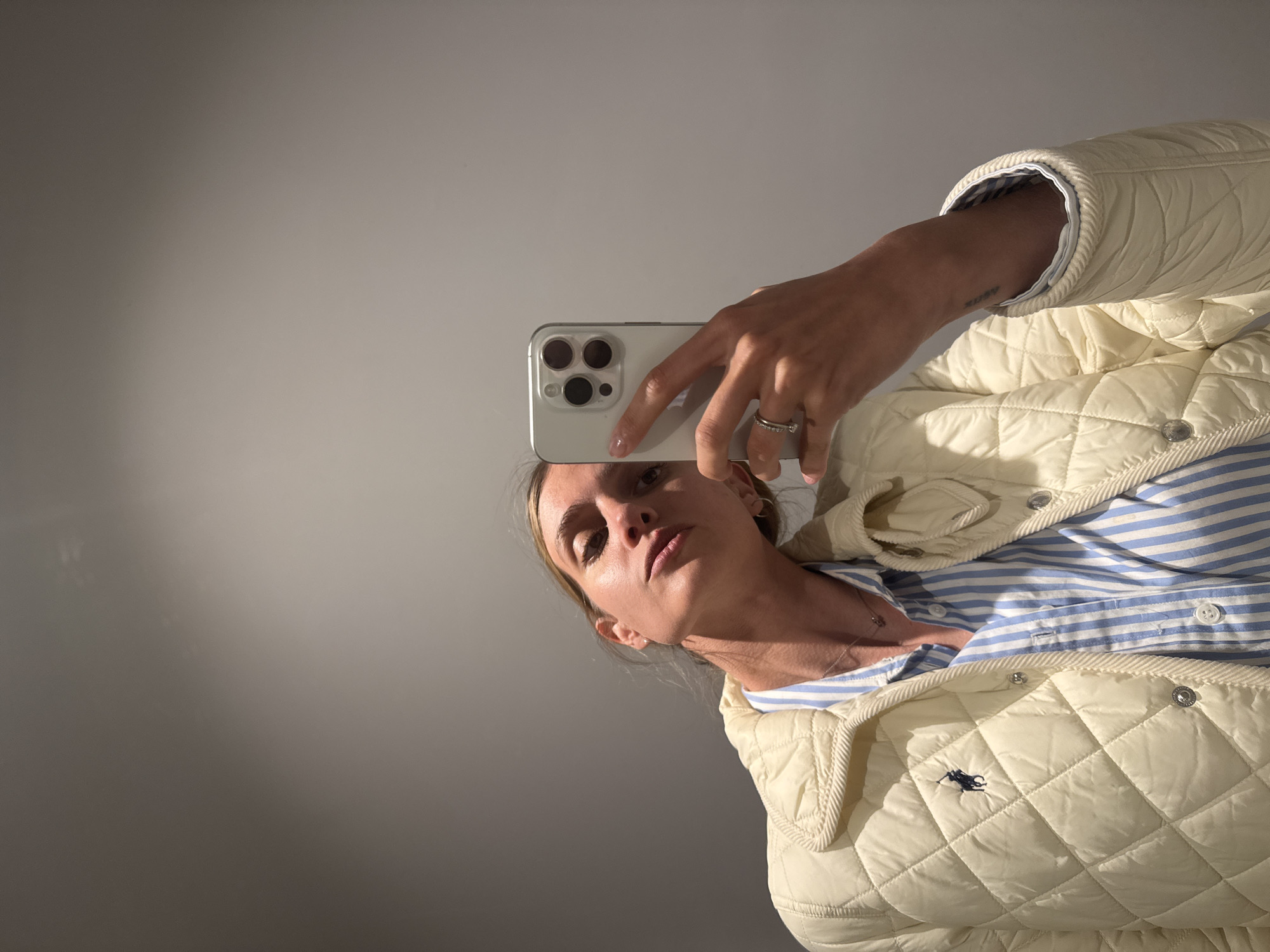 An upright portrait selfie, likely taken in a mirror, captures a person indoors in Tbilisi, Georgia. The subject, a person with light-toned skin, is partially visible from the forehead to the upper chest and right arm, looking directly into the camera with a neutral expression.
She is holding a silver smartphone, featuring a triple-lens camera module, in her right hand, which partially covers her face. A silver ring is worn on her right ring finger, and her fingernails have a light-colored polish. A faint text tattoo, possibly "RUBY," is visible on her inner right arm. Around her neck, a delicate necklace is visible.
The person is dressed in a light cream or off-white quilted jacket or vest, prominently displaying a small navy blue embroidered Polo logo (a horse and rider) on the left chest. The jacket features silver snap buttons. Beneath the jacket, a blue and white striped button-down shirt is worn.
The background consists of a plain, light-colored wall or ceiling. Soft, bright light illuminates the scene, casting a gradient effect from brighter areas at the top right to subtle shadows towards the bottom left. The overall setting appears to be a simple, well-lit interior.