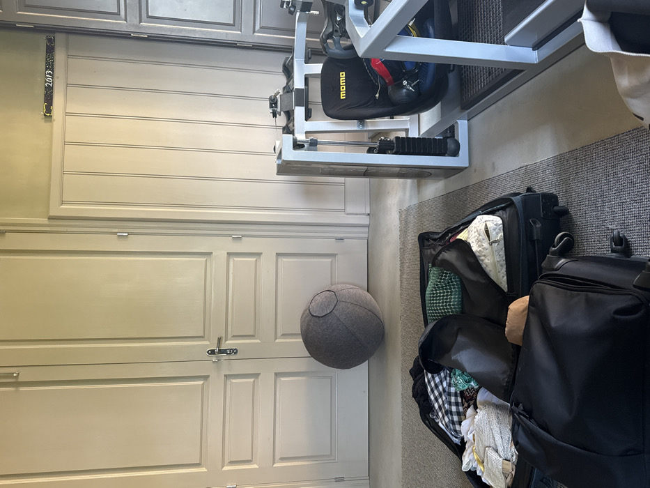 The image shows a light gray door with silver hardware, positioned beside a dark gray, round pouf resembling the Death Star. Adjacent to the pouf are two open black suitcases with clothing items inside. A silver exercise machine stands above the suitcases on a dark gray carpet. A decorative ribbon is seen above the door, possibly indicating a year.