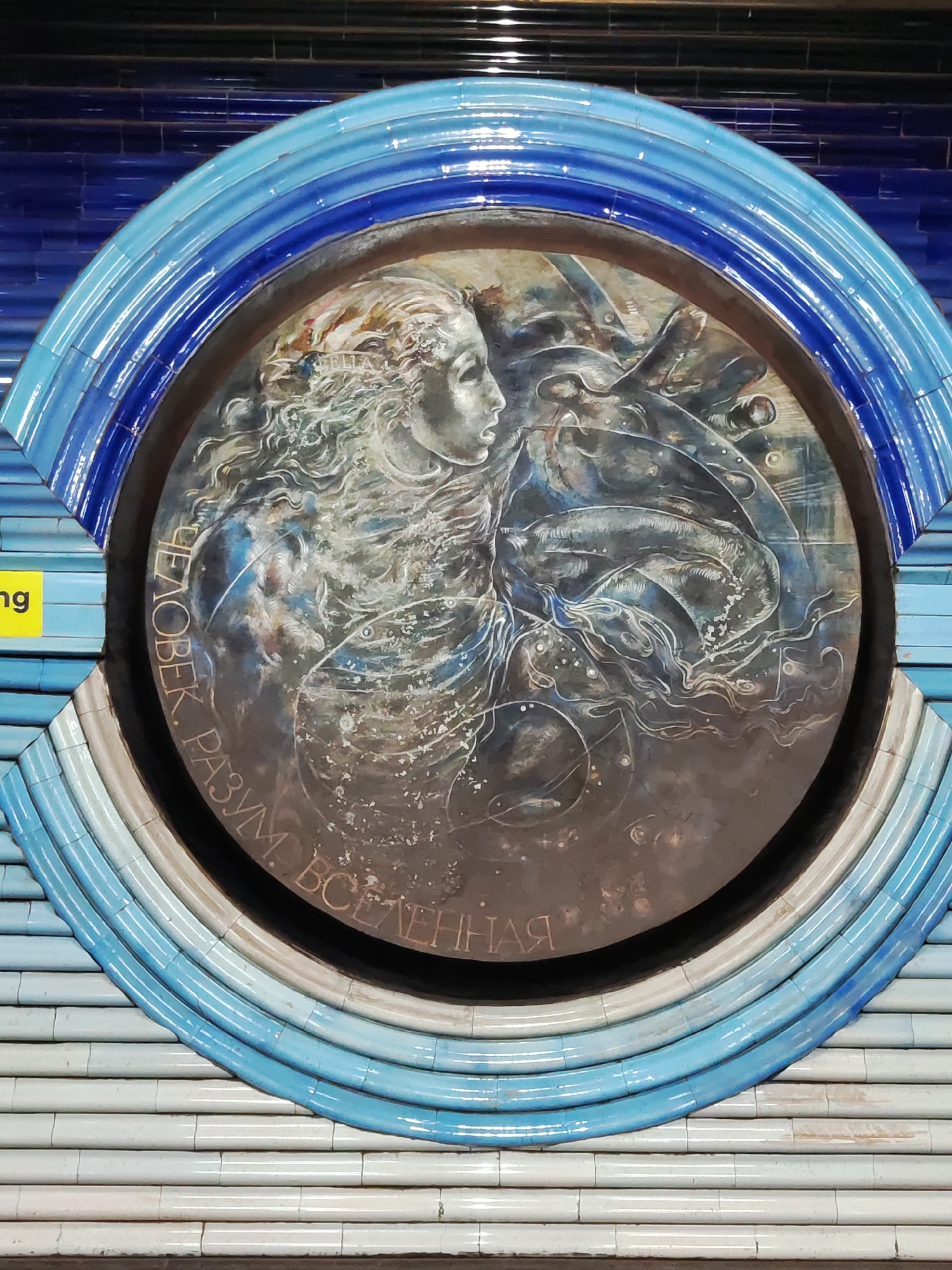 The image shows a circular mosaic artwork framed by blue and white ceramic tiles. The mosaic depicts a profile of a person's head and shoulders, with flowing hair, superimposed on a dark, swirling background that suggests cosmic elements like stars and nebulae. The artwork is framed by a series of concentric rings made of glazed ceramic tiles in varying shades of blue and white. These tiles are laid horizontally, creating a textured effect. The mosaic is mounted on a tiled wall, and some yellow signage is visible on the left side, partially obscured by the mosaic's frame. The overall impression is that of a public art installation, likely in a subway station or a similar public space, given the tiled environment. This specific mosaic is located at the Mirzo Ulugbek metro station in Tashkent, Uzbekistan, and is part of the station's design theme celebrating science and astronomy, with the inscription around the mosaic reading "Разум Человека Вселенная" which translates to "The Mind of Man is the Universe."