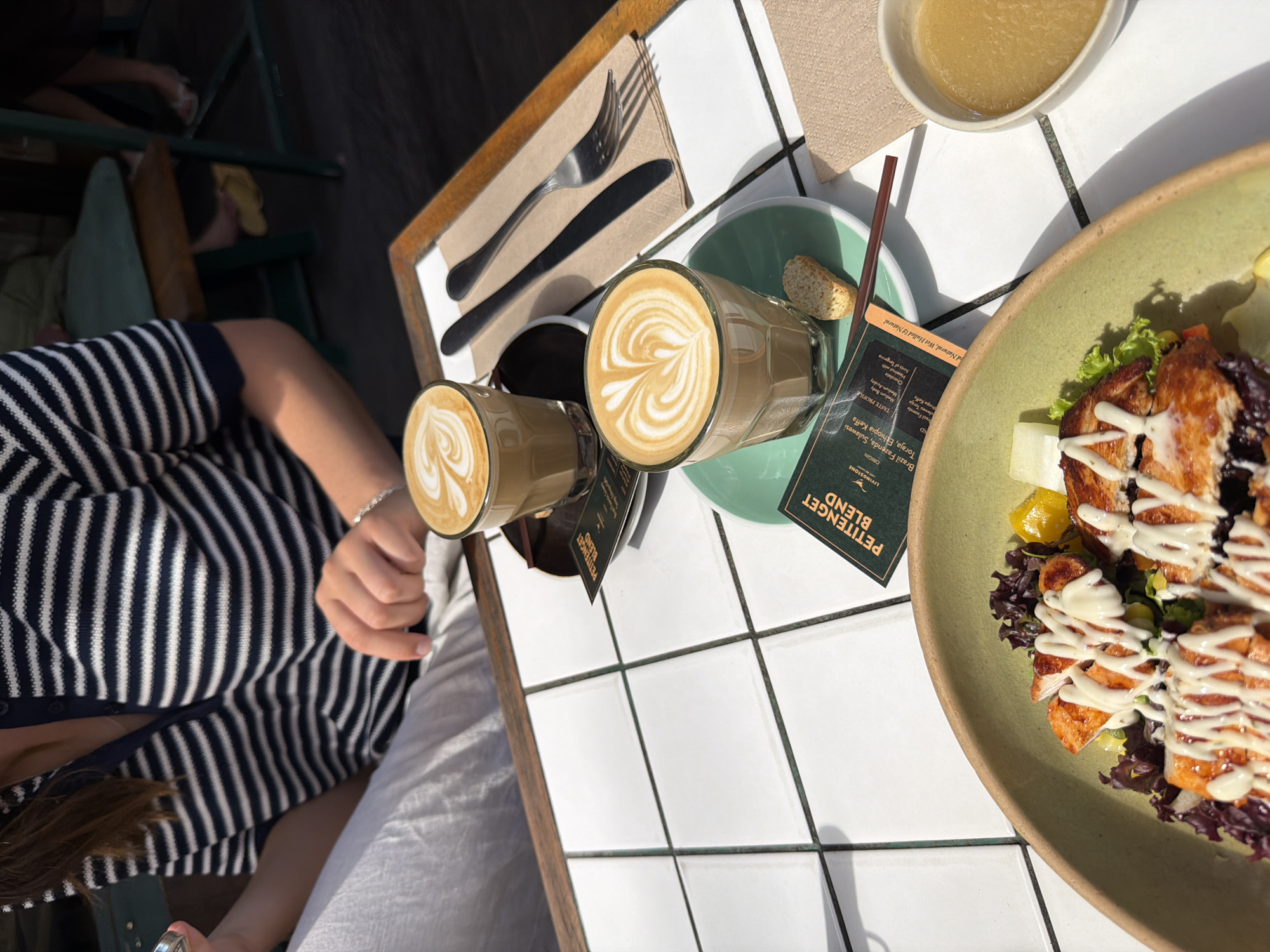 An overhead shot captures a sunlit outdoor cafe scene in North Kuta, Indonesia. A table with a white tiled surface is set for a meal. In the foreground, a green bowl contains a salad topped with grilled chicken and drizzled with white sauce, next to a small bowl of what appears to be sauce or dressing. Two glasses of coffee, each with latte art, are positioned towards the center. One glass sits on a green saucer, with a biscotti and a dark stirrer beside it. A menu card with "PETITENGET BLEND" visible is placed near the coffee. To the right of the coffees, a fork and knife rest on a light brown napkin. Partially visible in the background is a person wearing a navy and white striped polo shirt and white pants, with their hands resting on the table. Other patrons and cafe seating are blurred in the background.